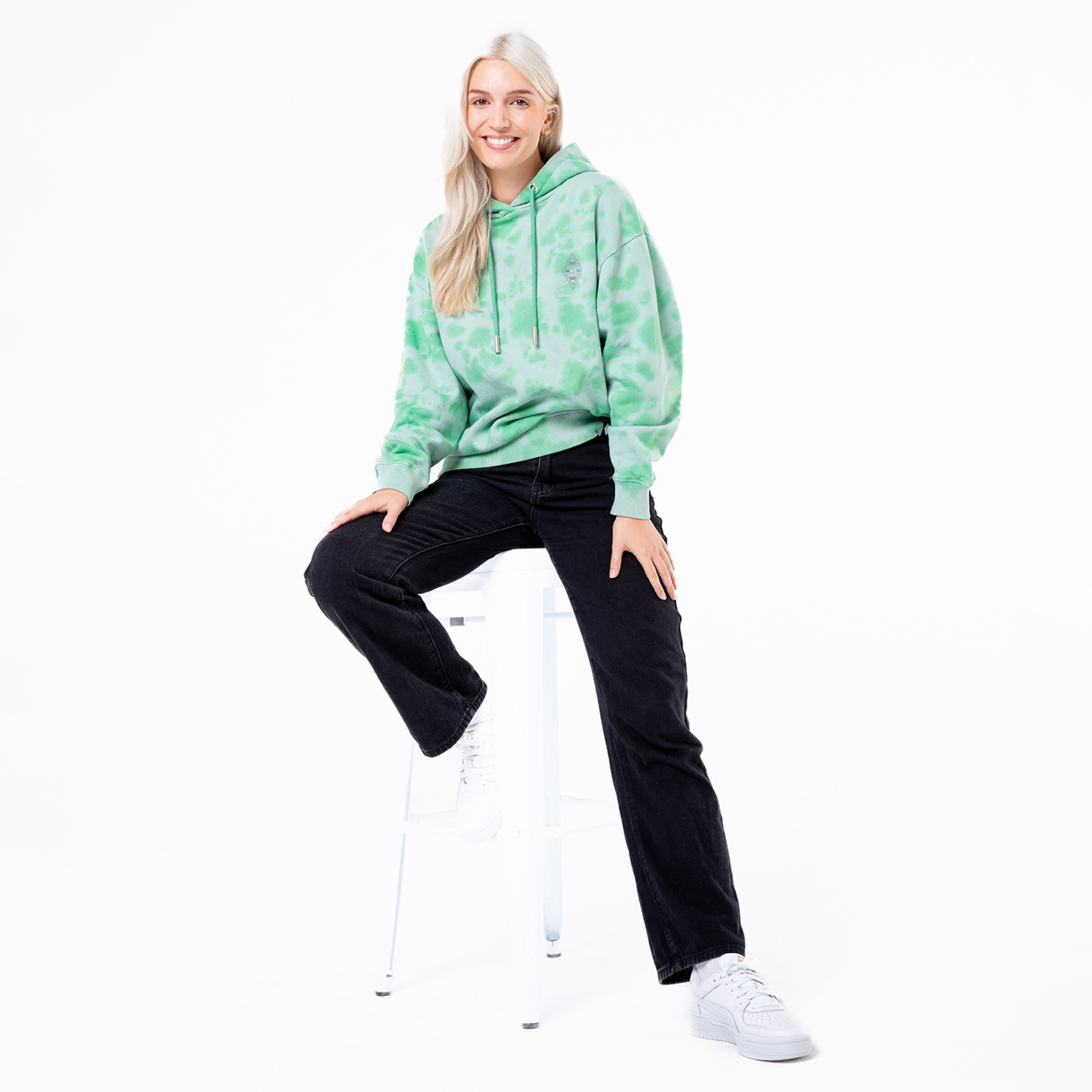 A woman sits on a white stool wearing a green patterned hoodie and black jeans.