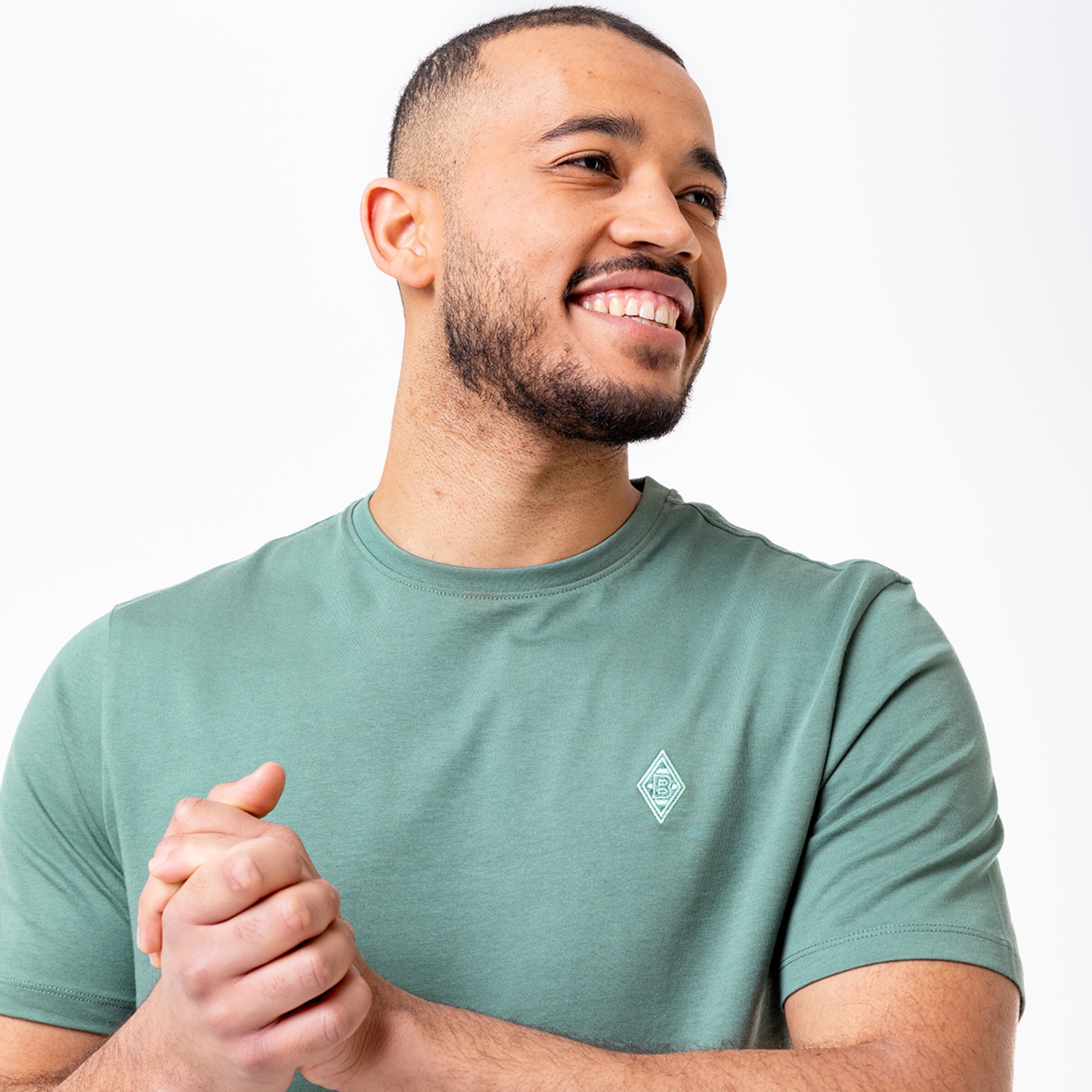 Man in green t-shirt with logo smiling and standing with clasped hands.