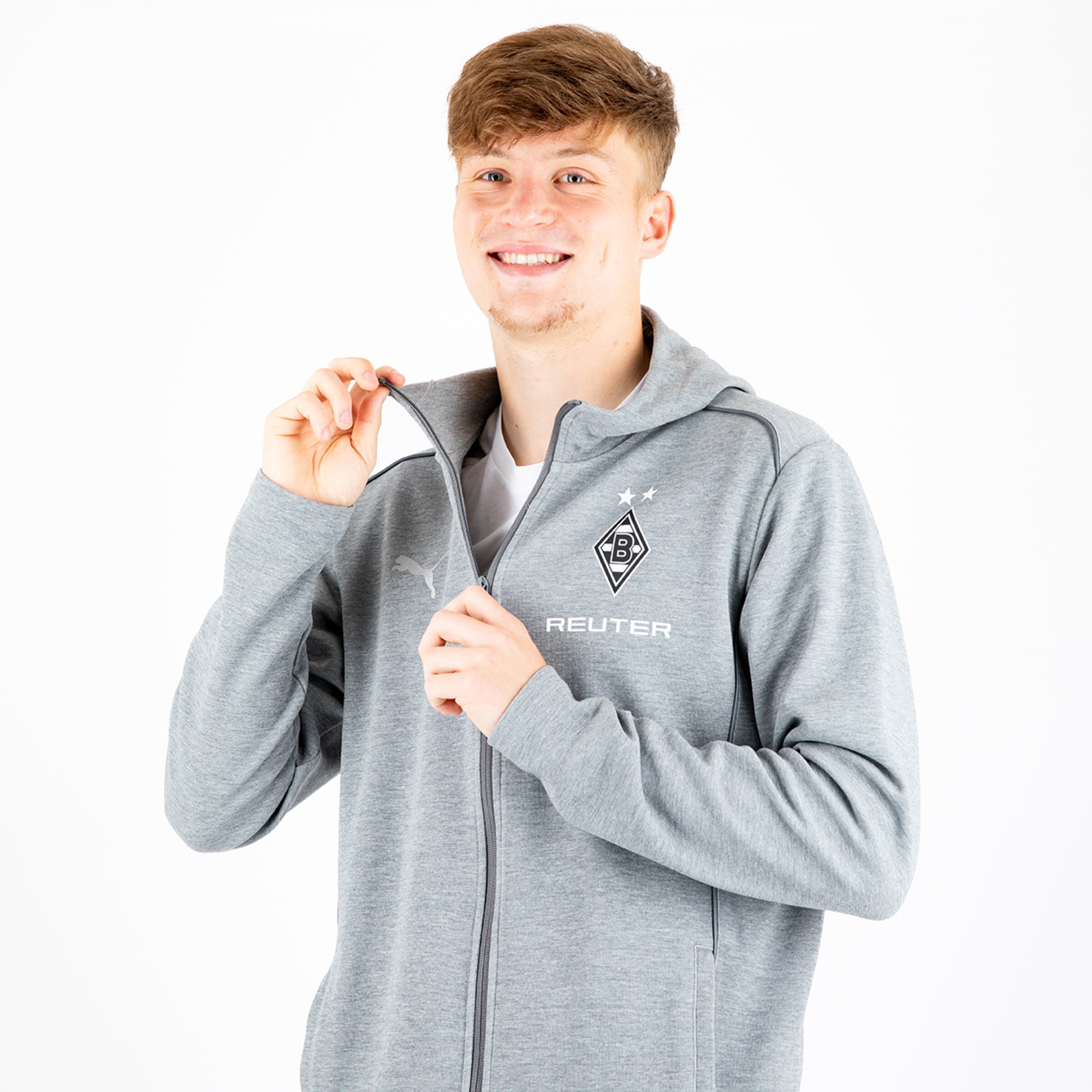 Ein junger Mann in einem grauen Sweatshirt mit Blitz-Logo der lächelt und seinen Reißverschluss leicht zieht.
