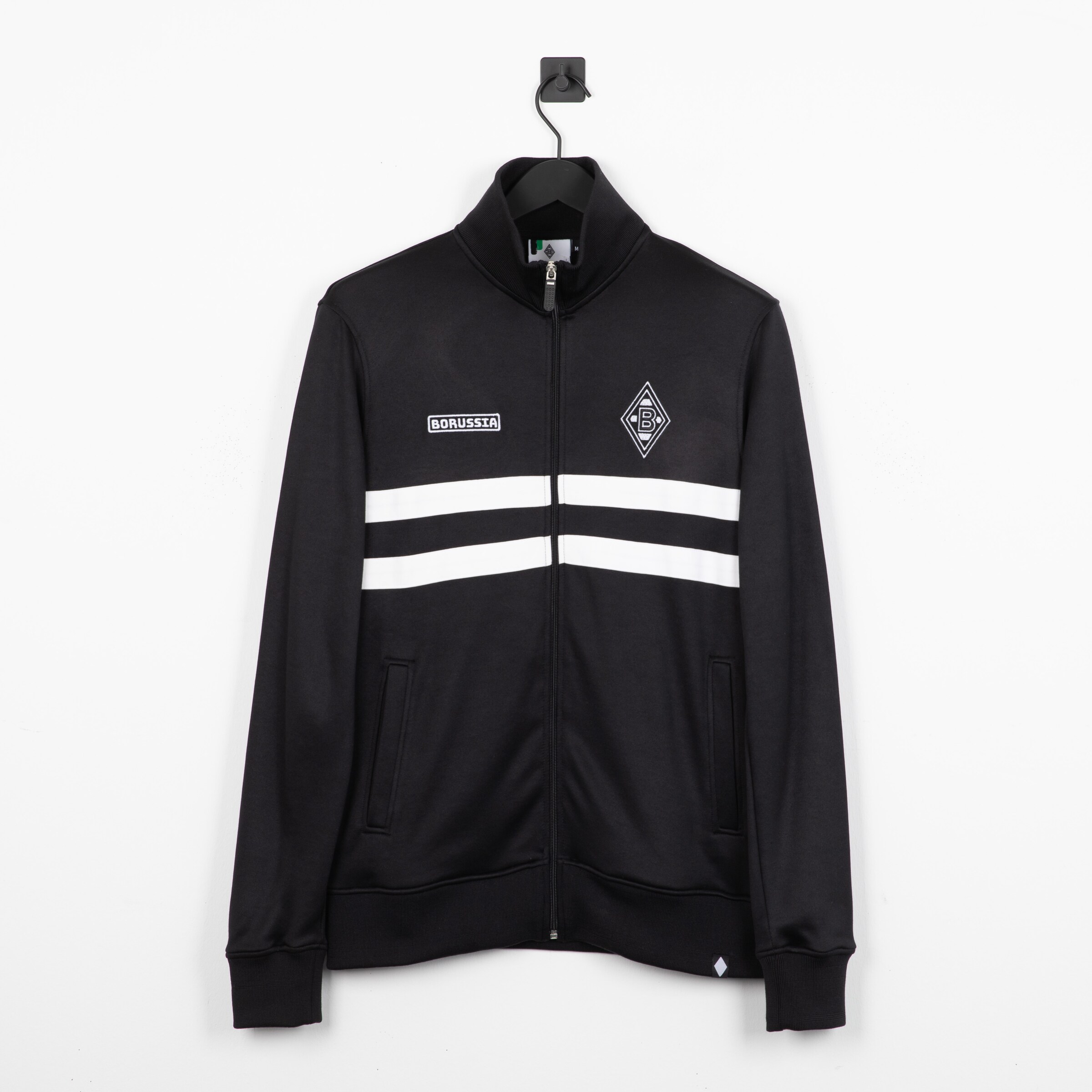 Schwarze Borussia Mönchengladbach Trainingsjacke mit zwei weißen Bruststreifen, Rauten-Logo links und Schriftzug rechts.