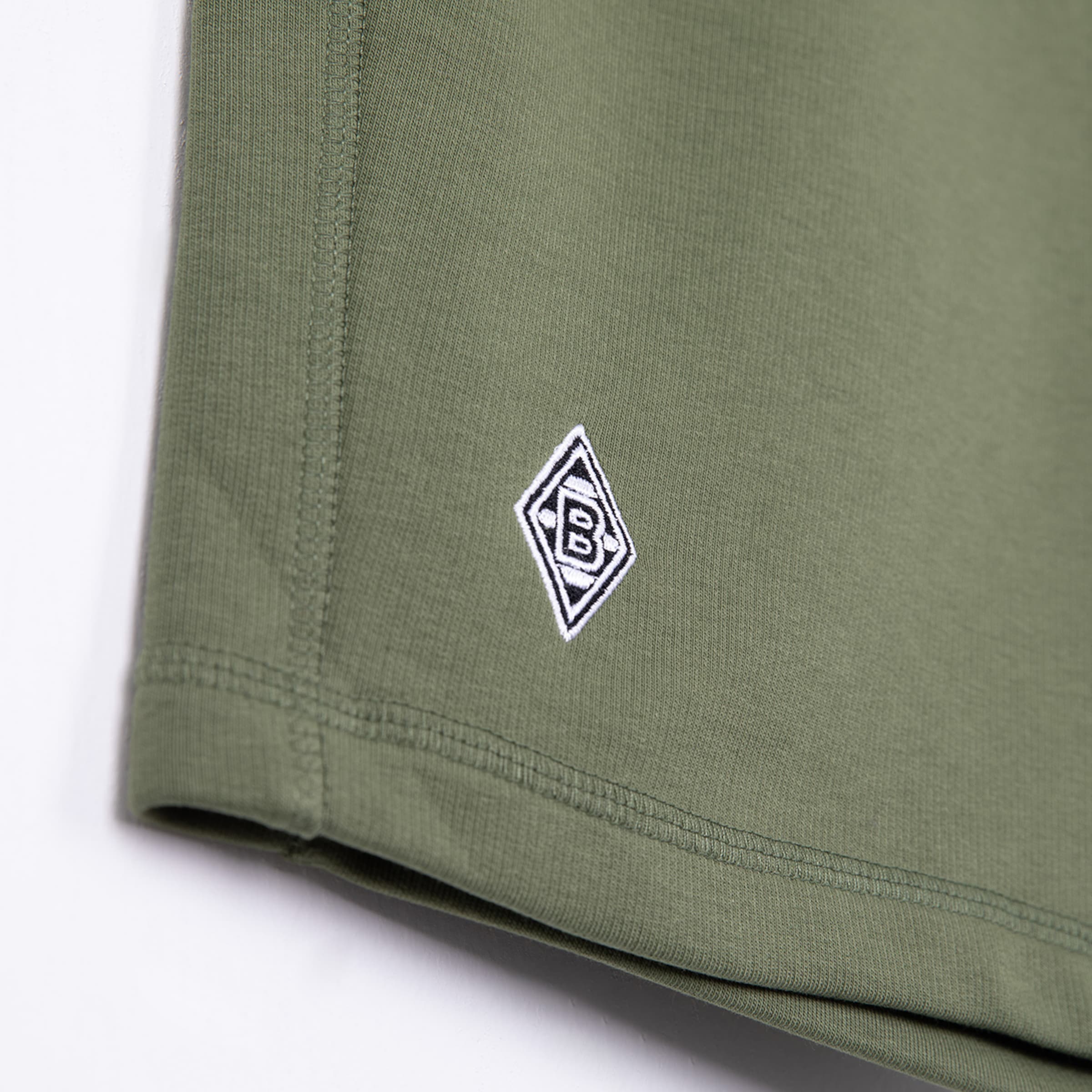 Nahaufnahme eines olivgrünen Kleidungsstücks mit dem eingestickten Rauten-Logo von Borussia Mönchengladbach.