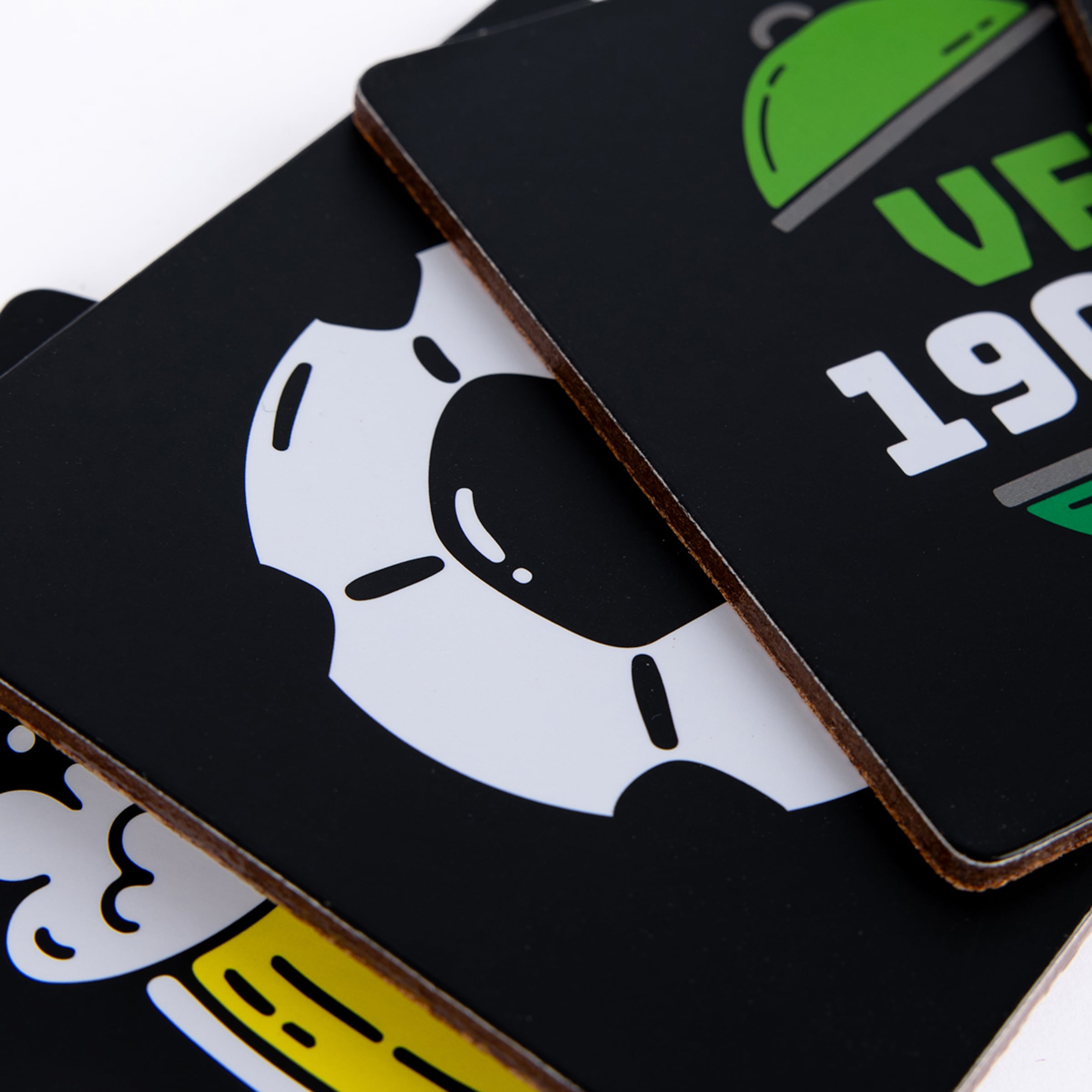 Nahaufnahme von drei bedruckten schwarzen Untersetzern mit bunten Grafiken und Korkseiten, gestapelt auf weißem Grund.