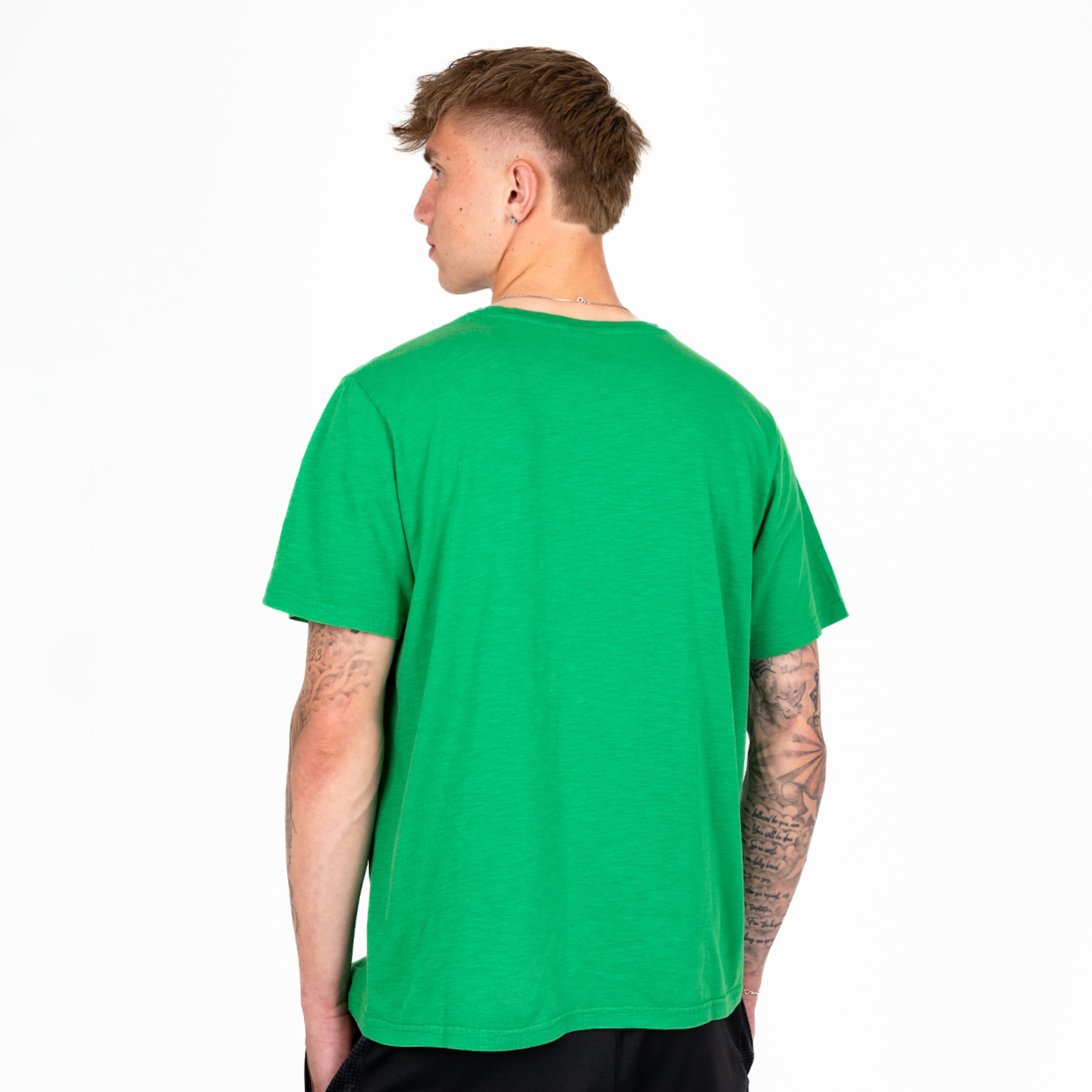 Rückenansicht eines Mannes in einem grünen T-Shirt und schwarzen Shorts vor weißem Hintergrund. Er hat tätowierte Arme.