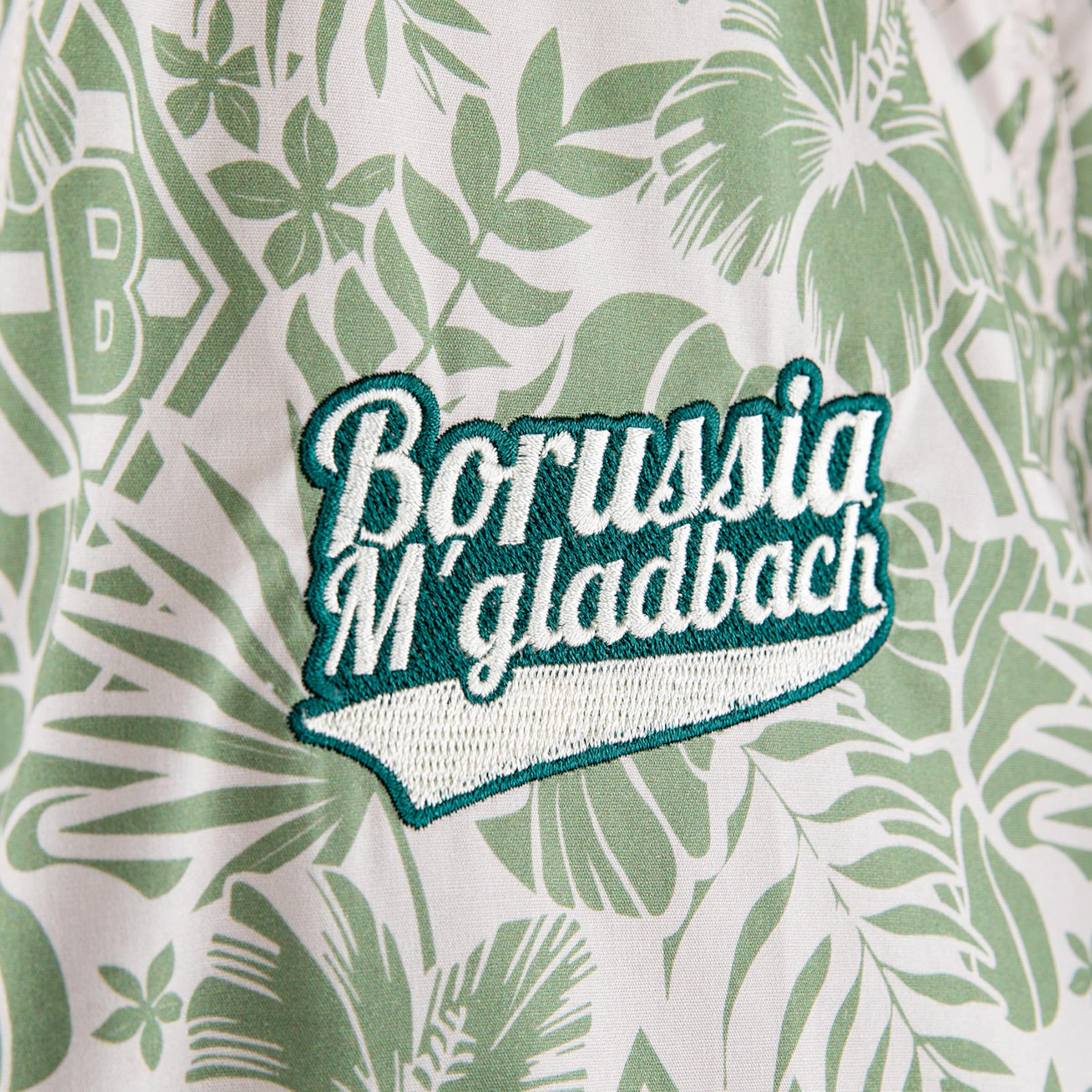 Nahaufnahme eines hellen Stoffes mit grünem tropischen Blattmuster. Zentral befindet sich ein gestickter Aufnäher mit 'Borussia M'gladbach' in weißer Schrift auf dunkelgrünem Hintergrund.