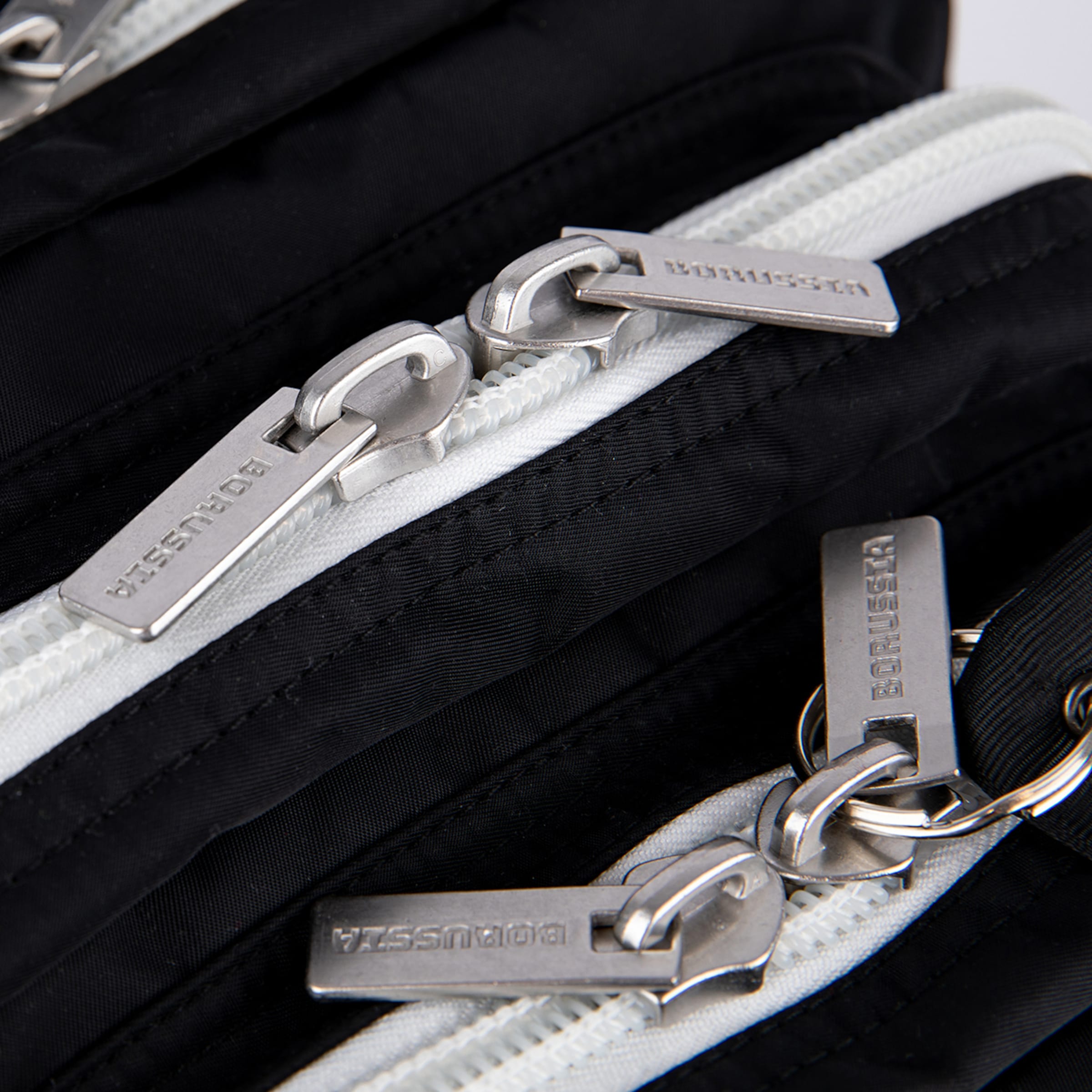 Close-up of a black bag with multiple white zippers and silver metal pulls. The zipper pulls are embossed with the word 'BORUSSIA'.