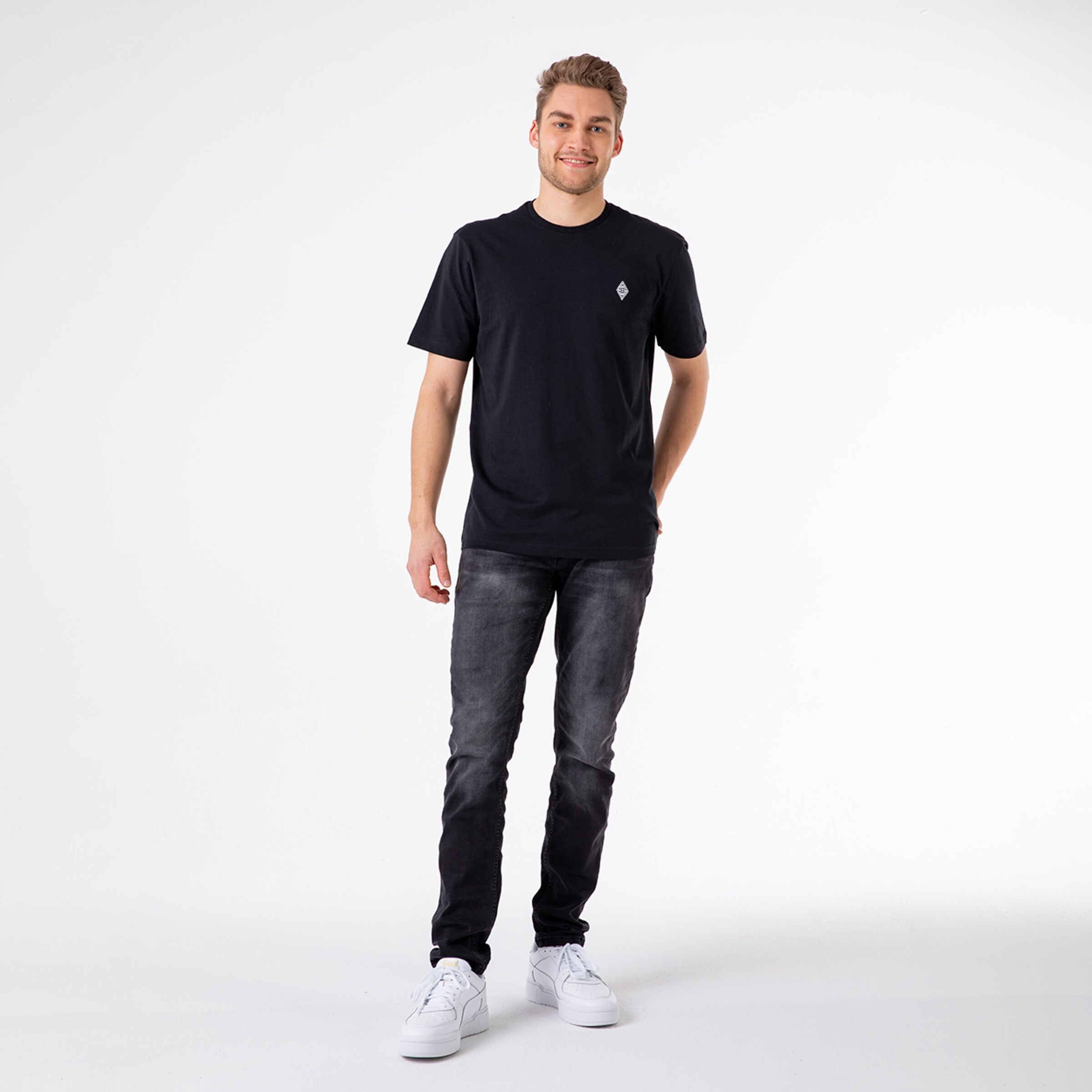 Young man in a black T-shirt and jeans standing with one hand in his pocket.