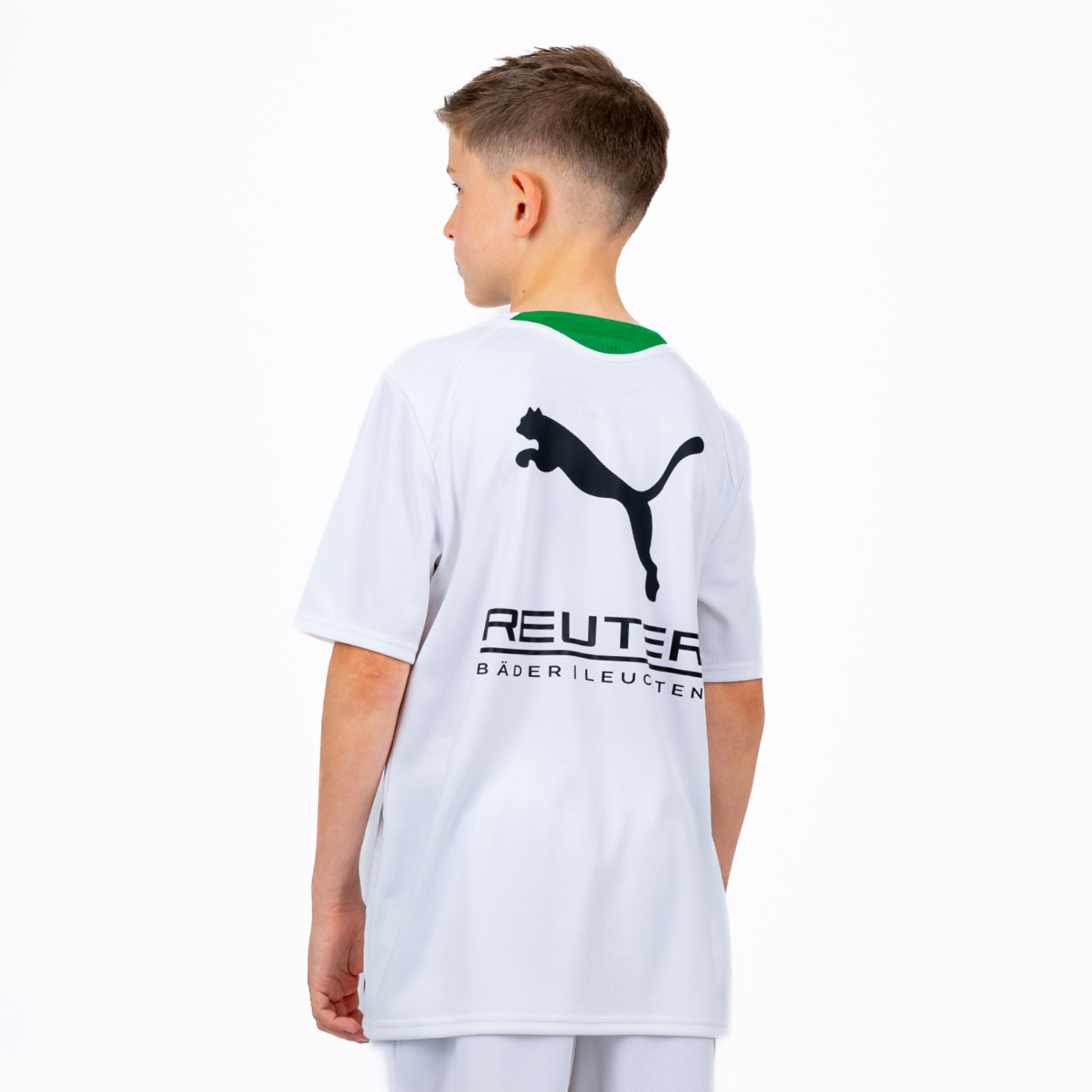 A young boy, viewed from the back and slightly turned to his left, wears a white t-shirt with a green collar. On the back of the shirt, there is a large black Puma logo, with the text "REUTER" and "BÄDER | LEUCHTEN" printed below it.