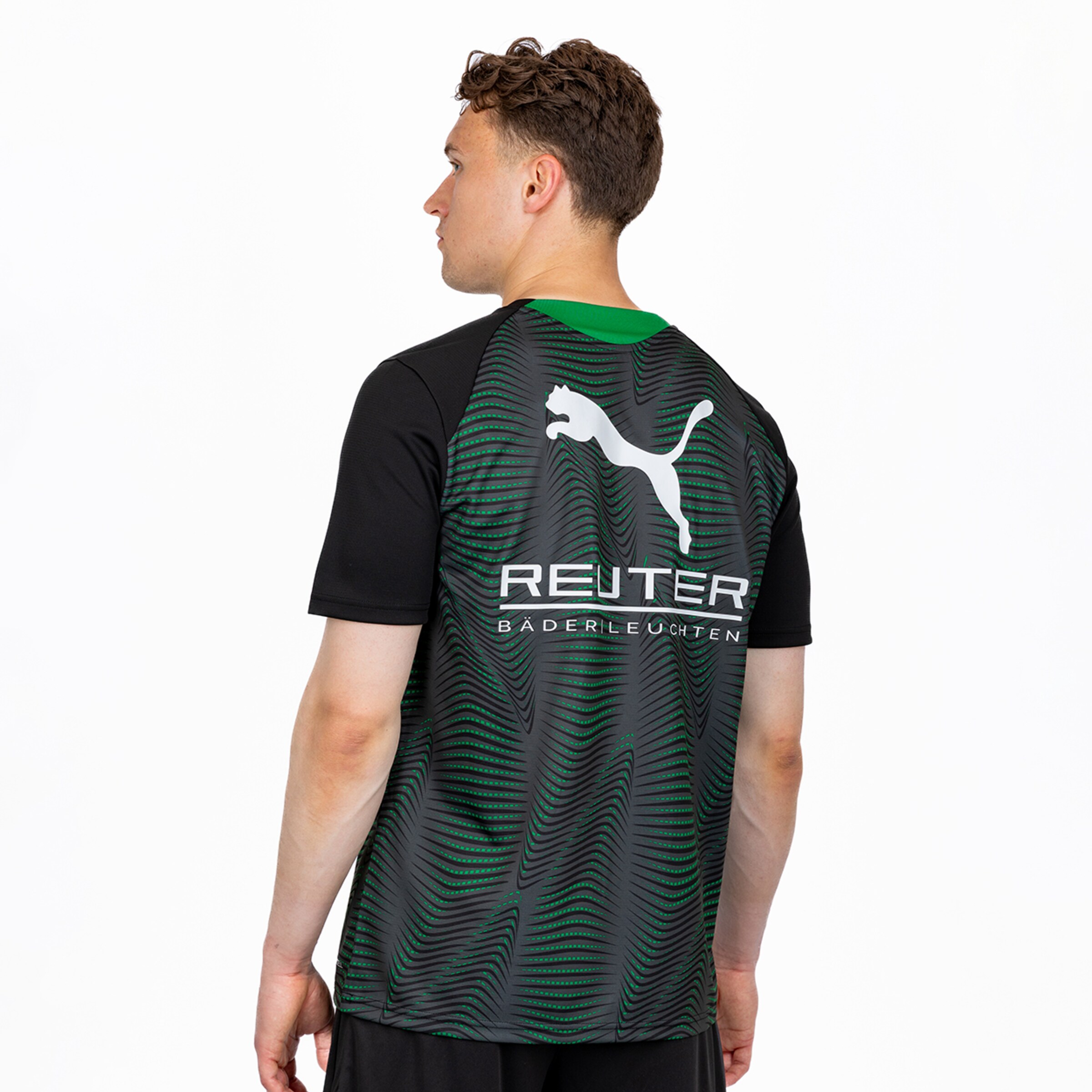 Rückenansicht eines Mannes in einem schwarz-grau-grün gemusterten T-Shirt mit weißem Puma-Logo und dem Text „REUTER BÄDERLEUCHTEN“ auf weißem Hintergrund.