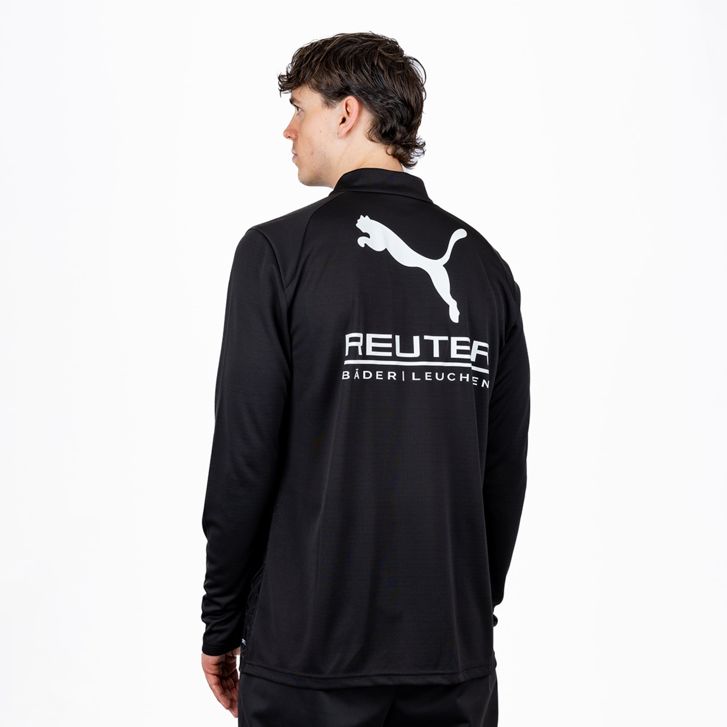 Ein Mann von hinten, leicht nach links gedreht, vor einem weißen Hintergrund. Er trägt eine schwarze, langärmelige Sportjacke mit einem weißen Puma-Logo und dem Schriftzug „REUTER BÄDER | LEUCHTEN“ auf dem Rücken. Er hat dunkles, welliges Haar.