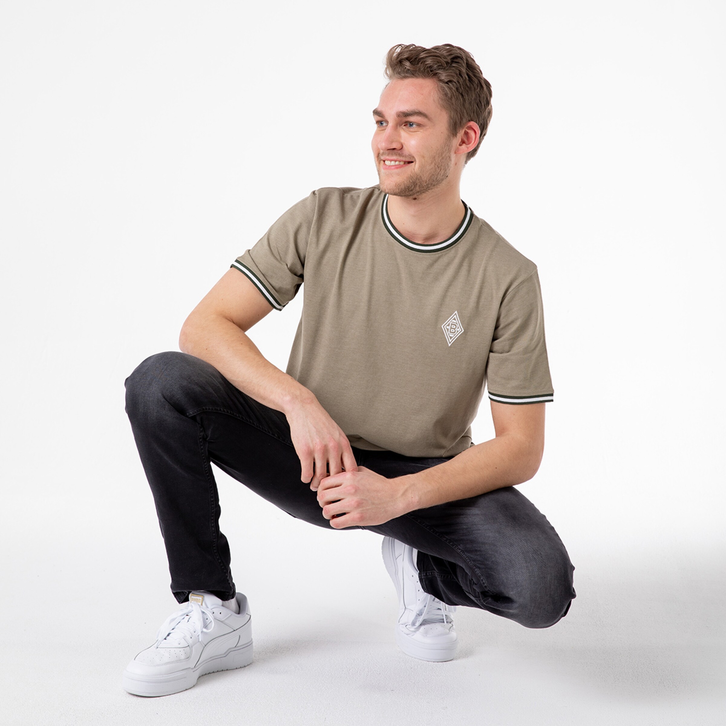 Man in khaki t-shirt with green stripes and black jeans crouching and smiling.