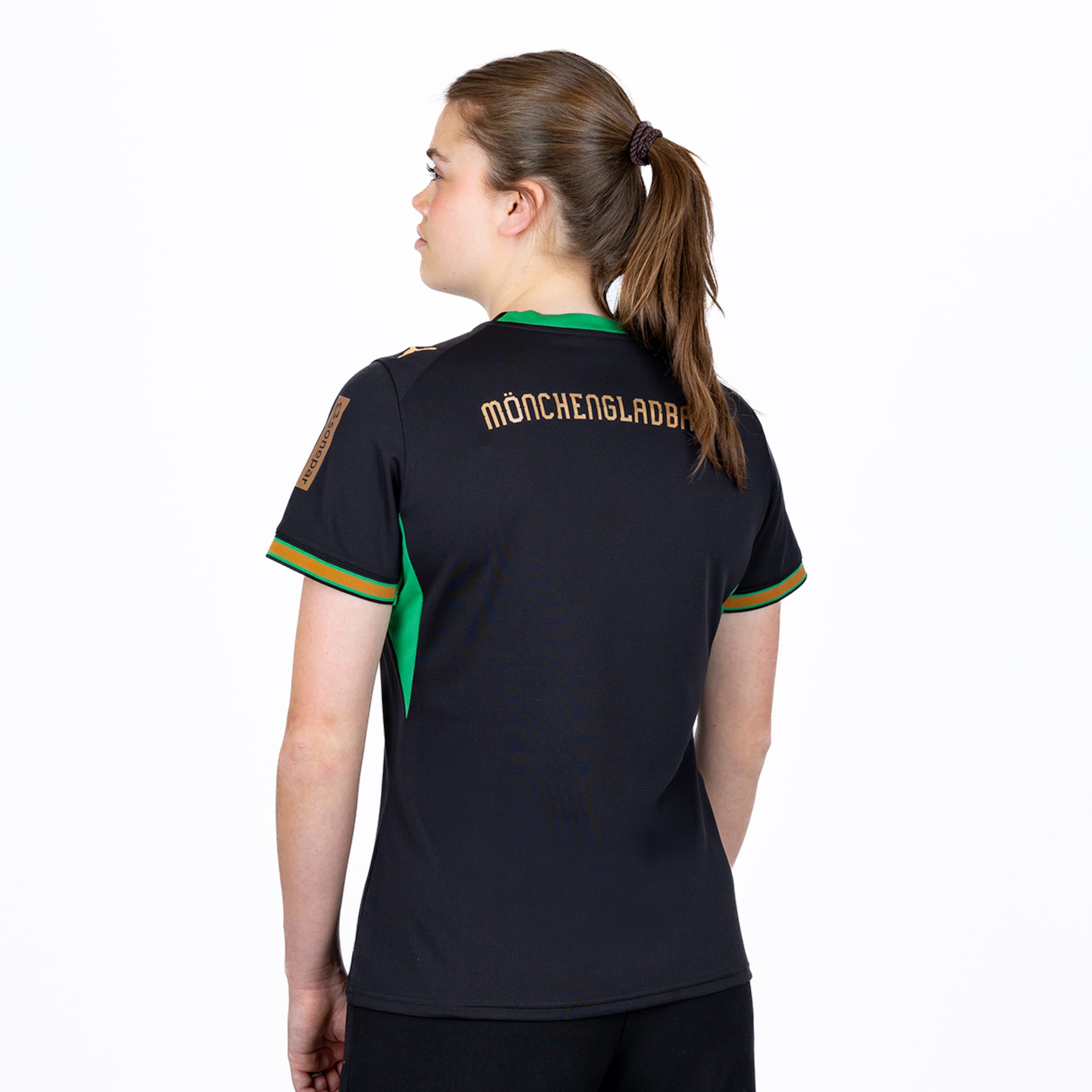 Rückansicht einer Frau in einem schwarzen Mönchengladbach-Trikot mit grünen und goldenen Details vor weißem Hintergrund.
