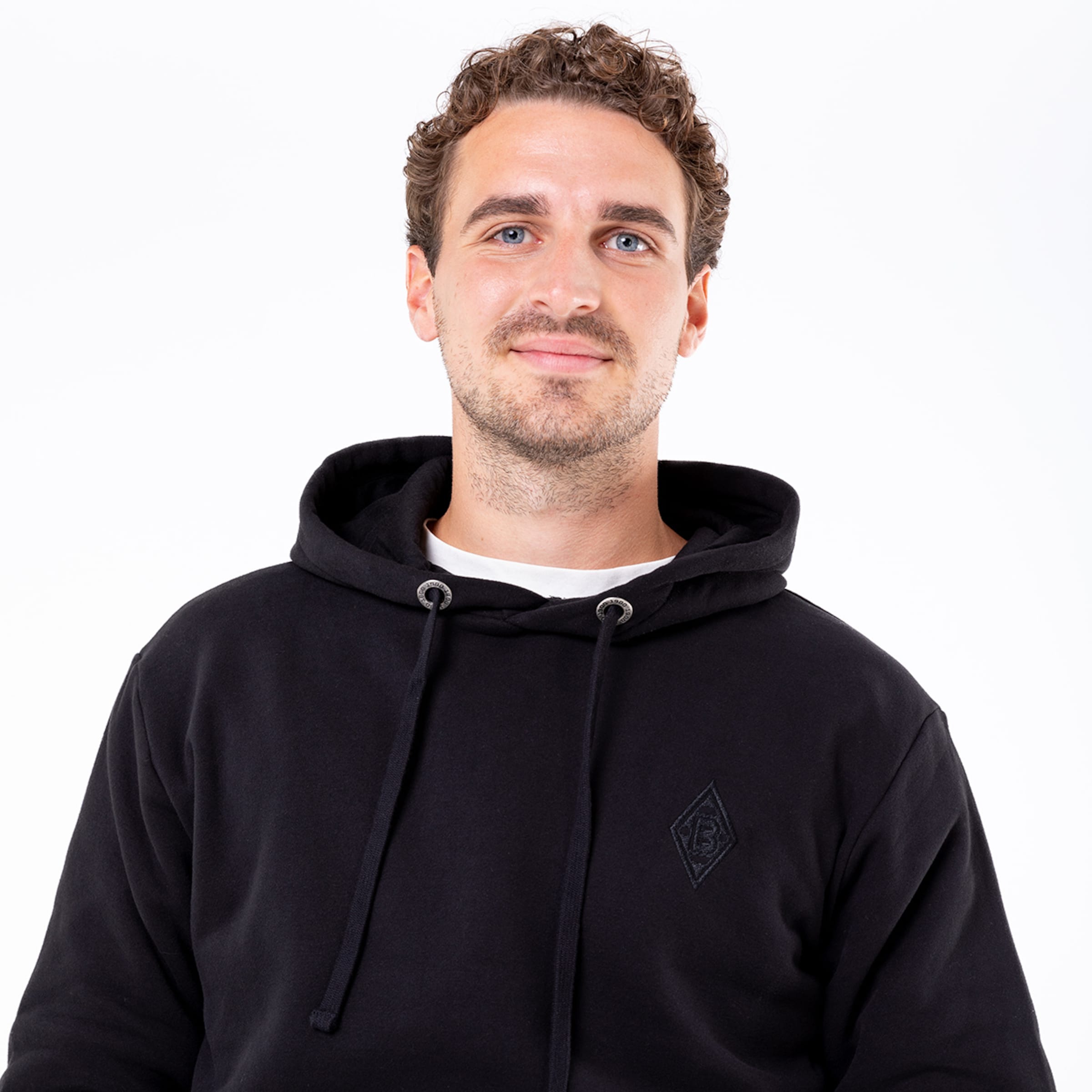 Ein junger Mann in einem schwarzen Hoodie mit Kapuze steht vor neutralem Hintergrund und lächelt leicht.