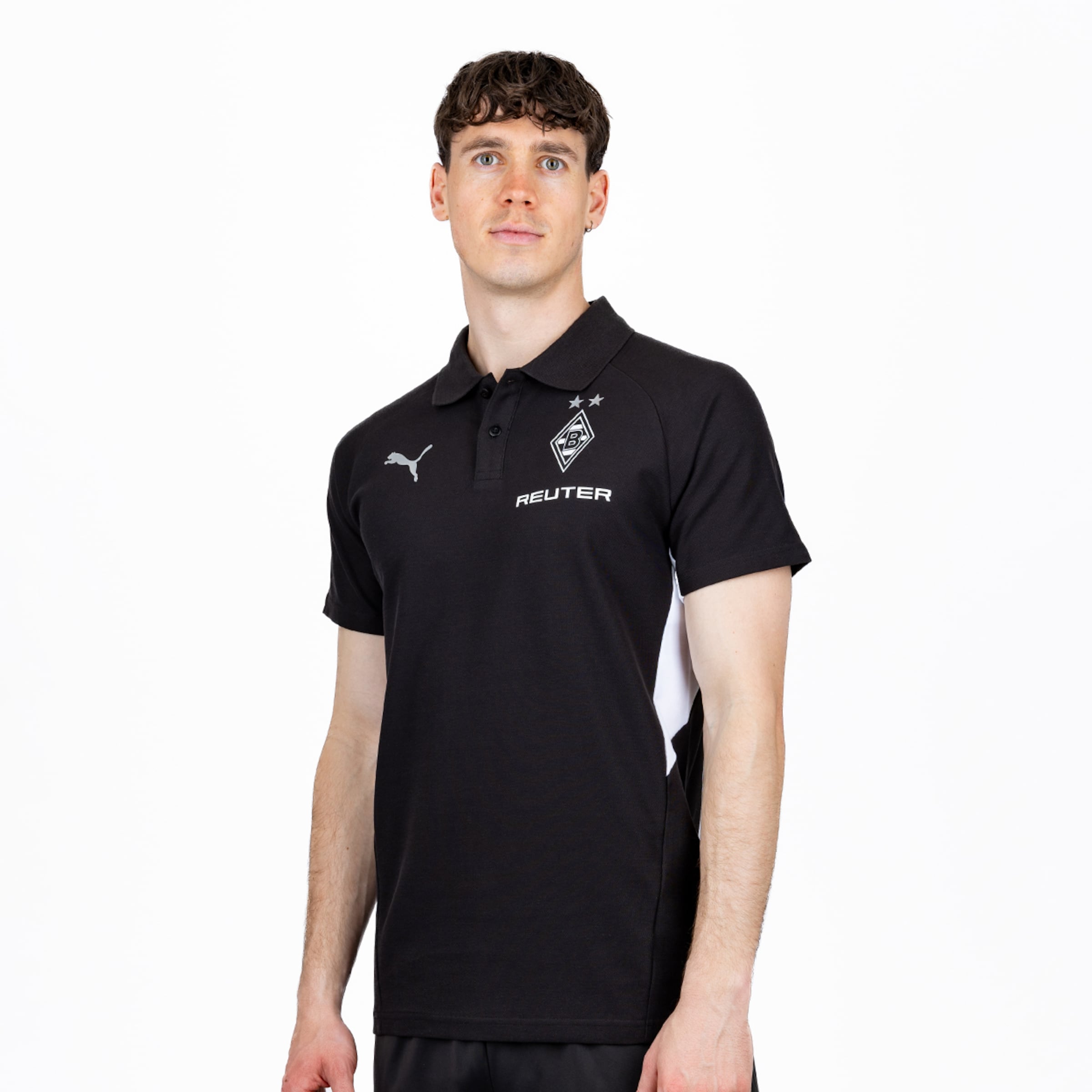 Ein hellhäutiger Mann mit dunklen, lockigen Haaren und hellen Augen trägt ein schwarzes Poloshirt mit Puma-Logo, Borussia Mönchengladbach-Wappen und „REUTER“-Schriftzug. Er blickt nach vorne.