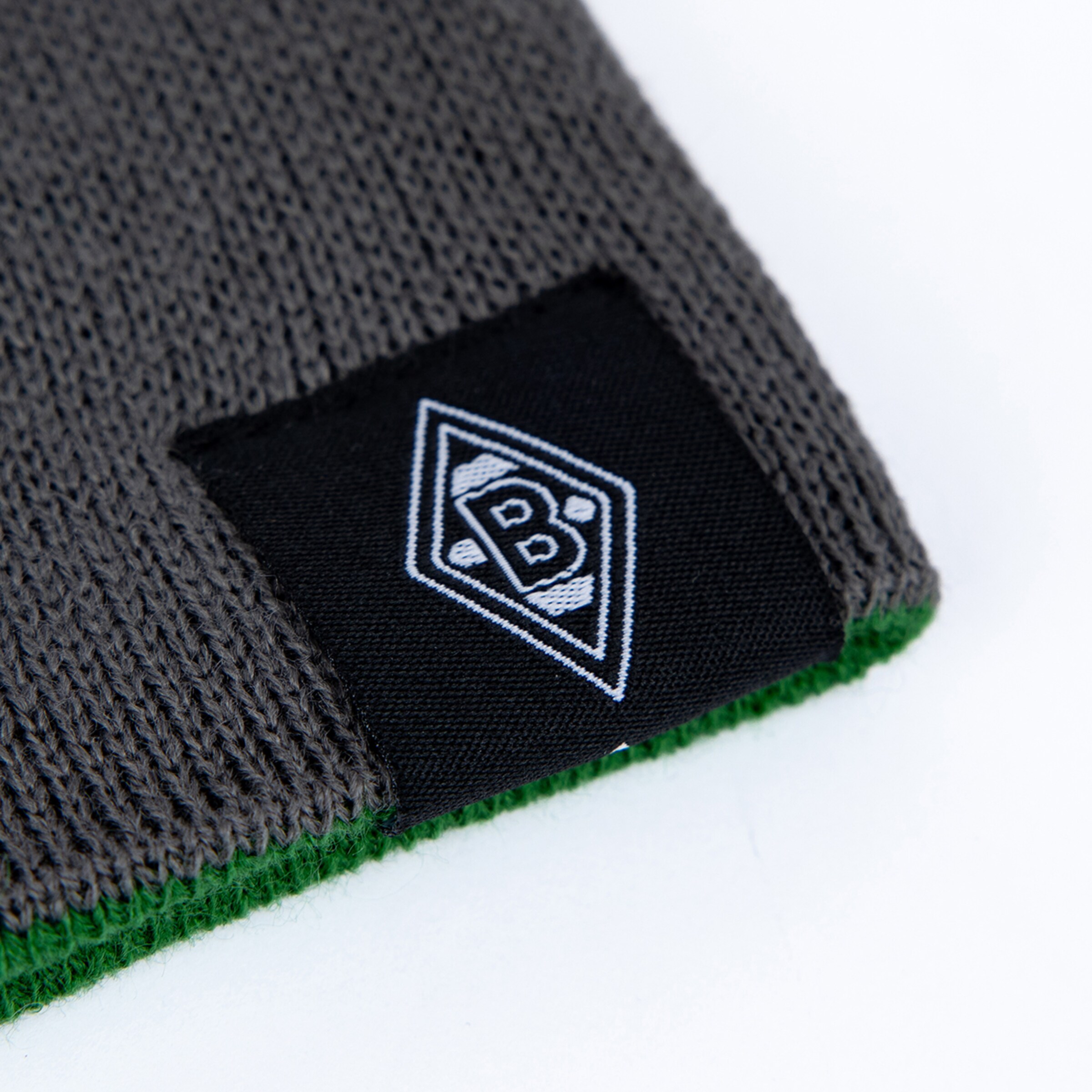 Nahaufnahme einer grauen Strickmütze mit grünem Rand und schwarzem Patch, der das weiße Borussia Mönchengladbach Logo (Raute mit B) zeigt.
