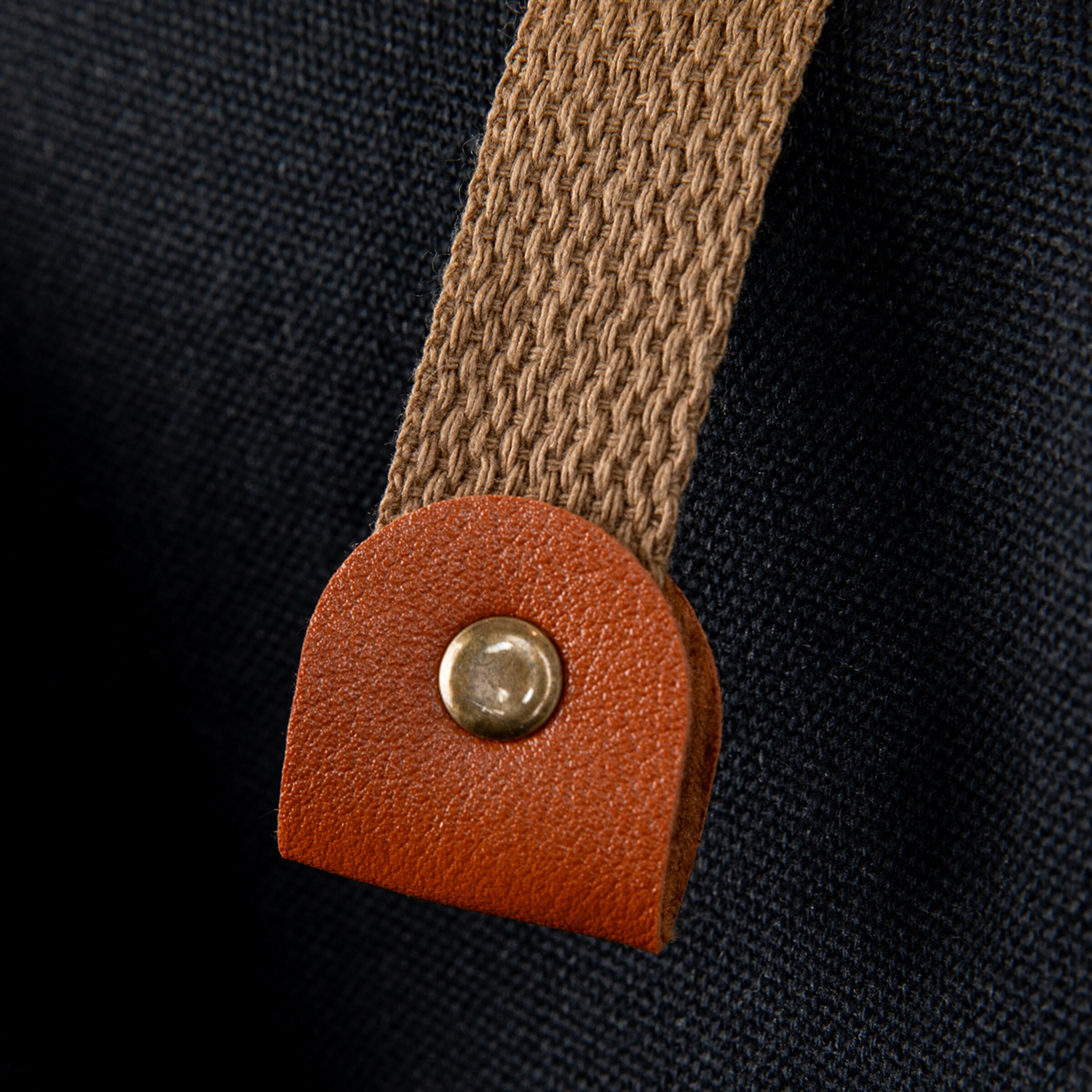 Close-up of a textured brown woven strap attached to a dark fabric with a small, curved brown leather tab and a brass-colored rivet. The textures of the fabric, strap, and leather are clearly visible.