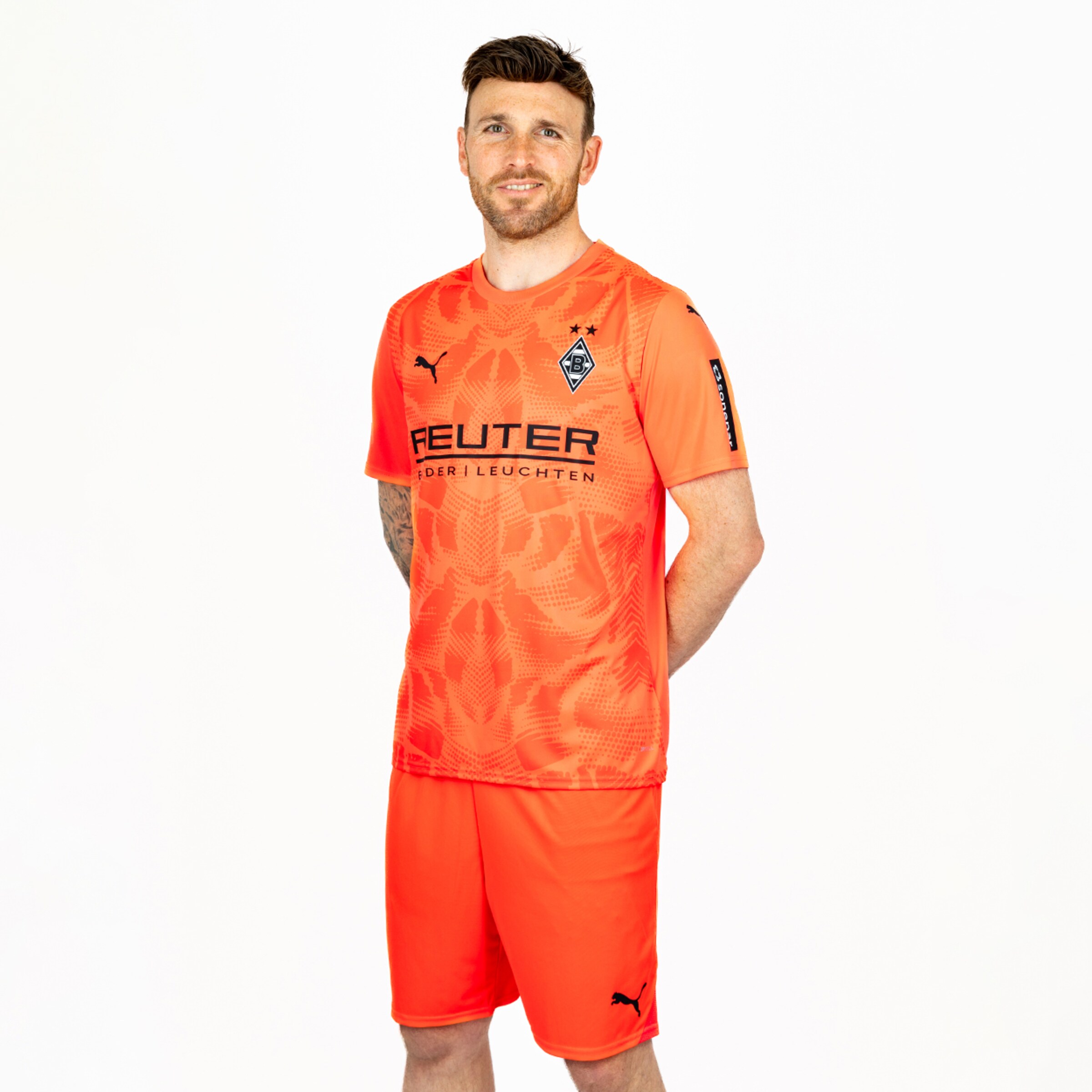 Football player in an orange Mönchengladbach kit and shorts against a white background. Puma and Reuter logos are visible.
