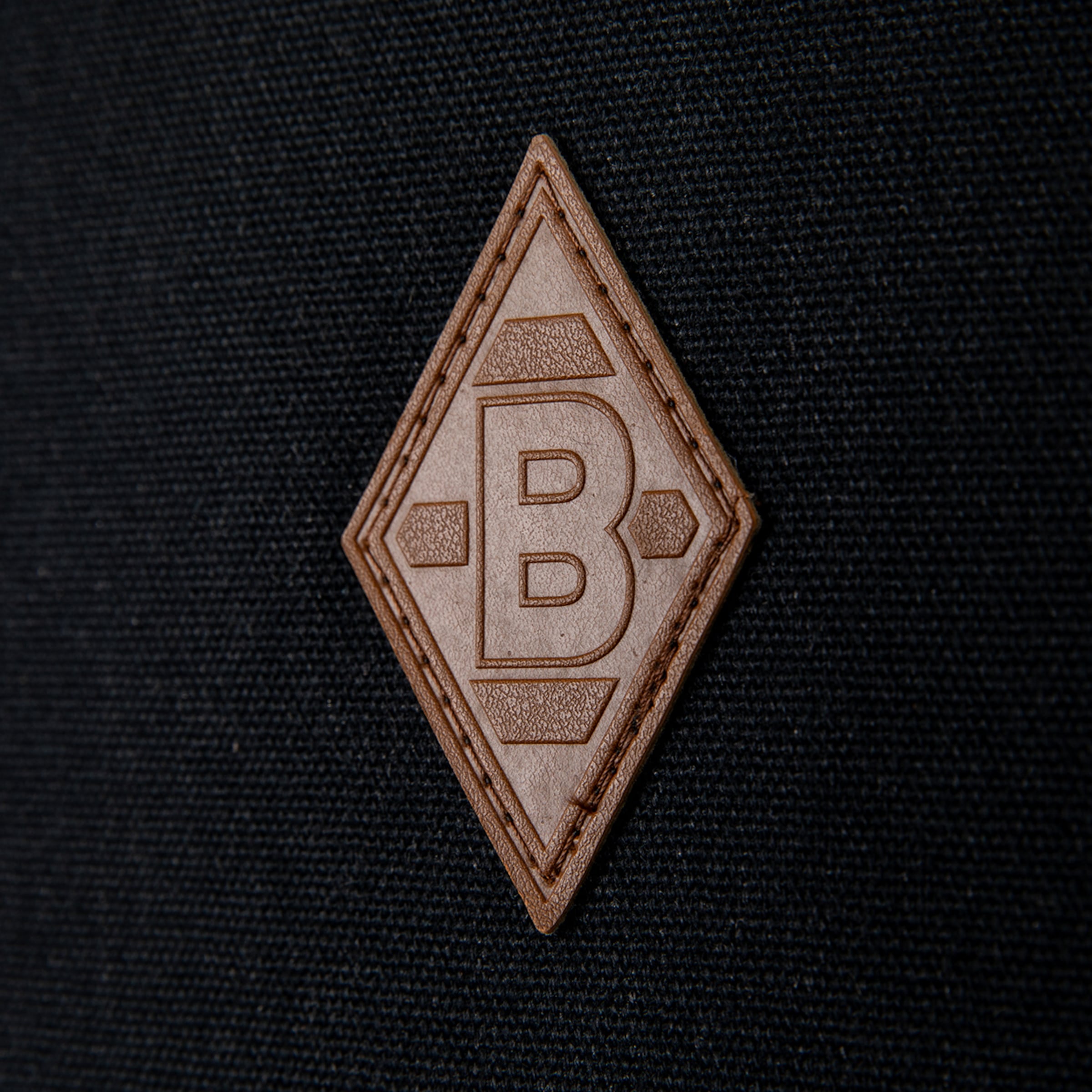 Close-up of a brown, diamond-shaped leather patch featuring an embossed letter 'B' and geometric designs, stitched onto a dark, woven fabric background.