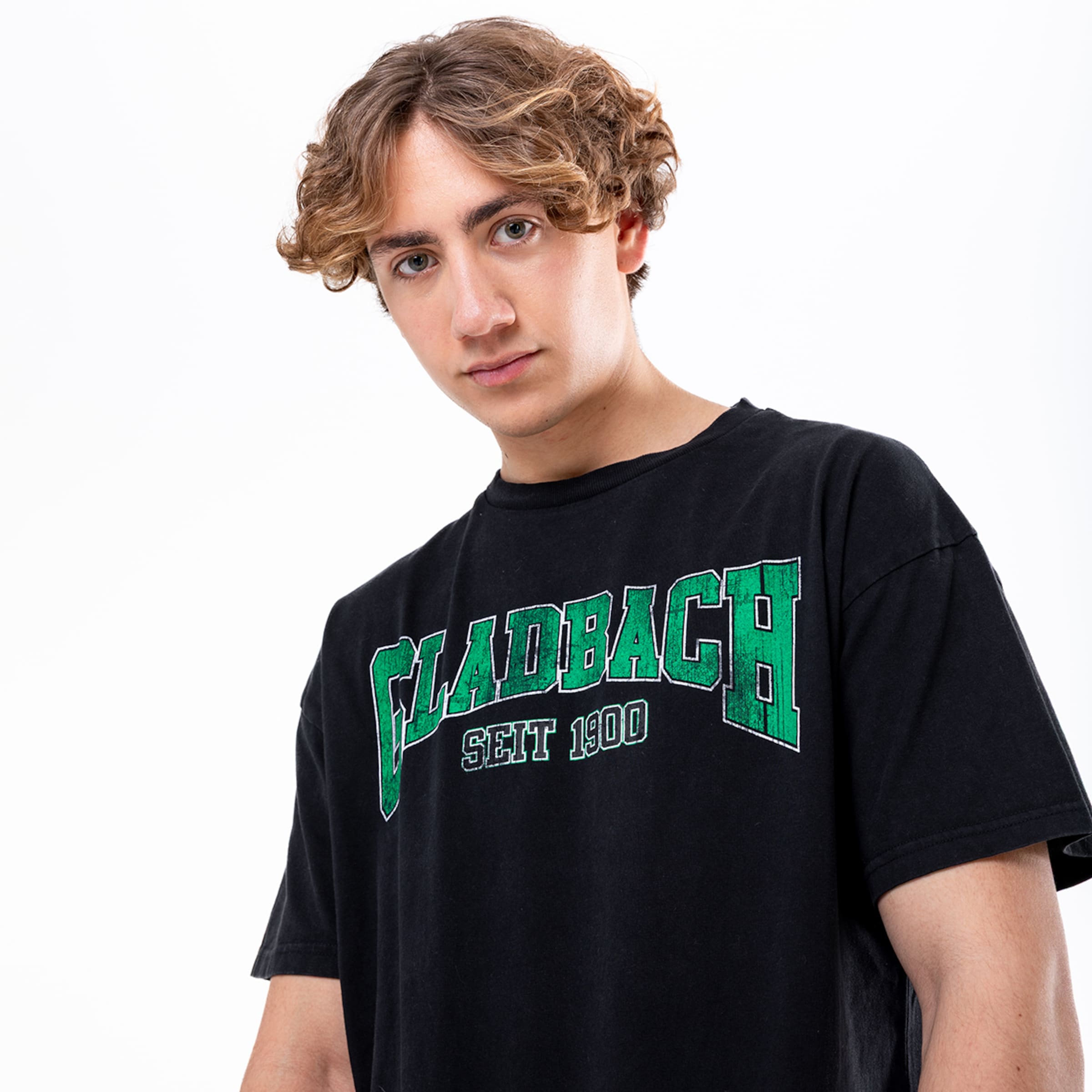Junger Mann mit lockigem Haar im schwarzen T-Shirt mit grünem GLADBACH SEIT 1900 Aufdruck vor weißem Hintergrund.