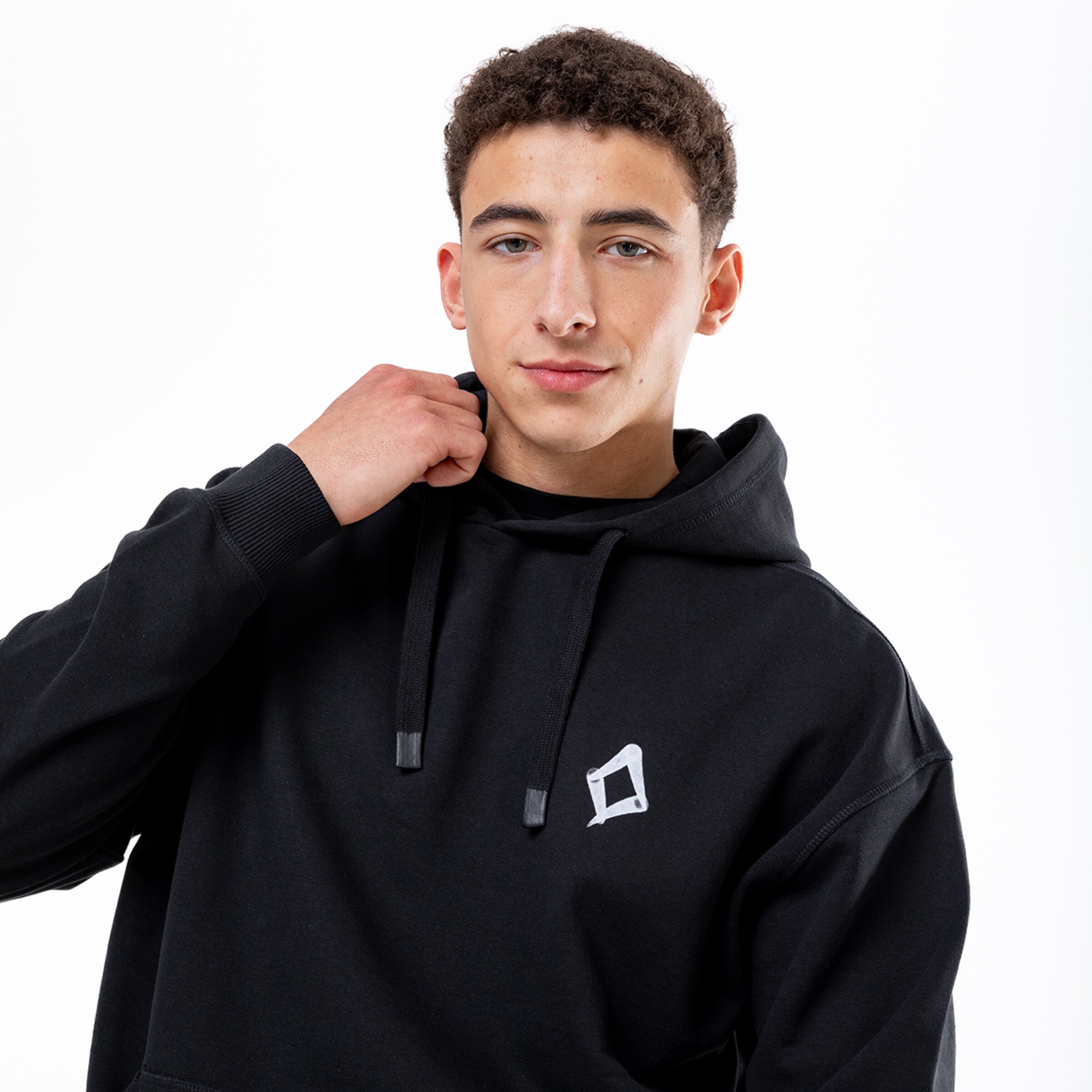 Junger Mann mit braunen Locken im schwarzen Hoodie mit weißem Logo, posiert vor weißem Hintergrund und berührt seinen Kragen.