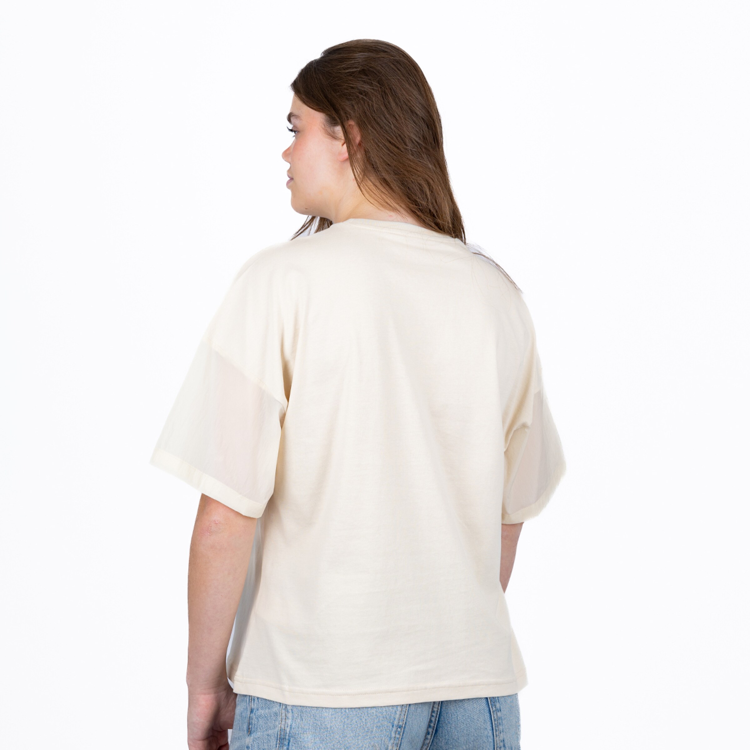 Rückansicht einer Frau mit langen braunen Haaren in einem cremefarbenen Oversized-T-Shirt und hellblauen Jeans vor weißem Hintergrund.