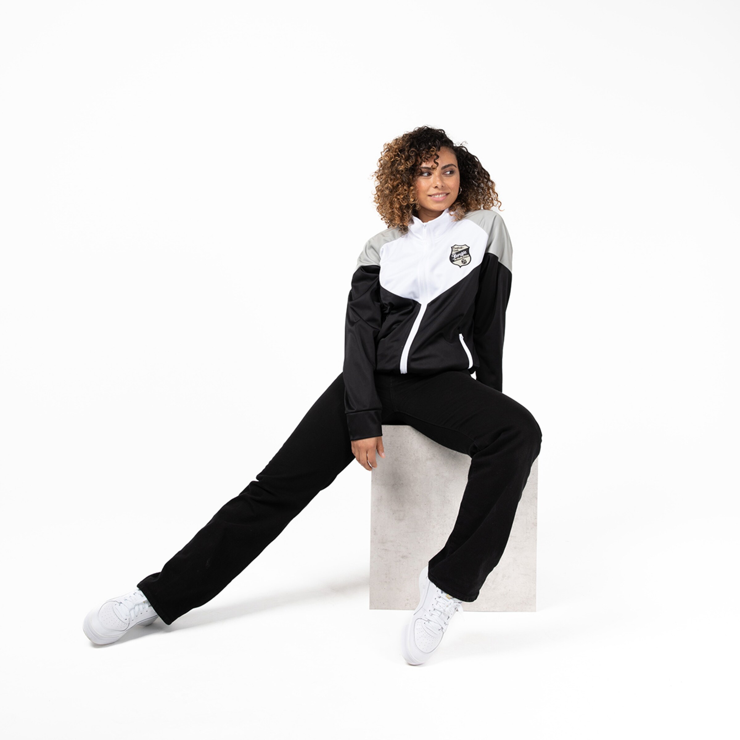 Eine Frau in einem sportlichen Outfit mit einer Jacke und einer Hose sitzt entspannt auf einem Block.