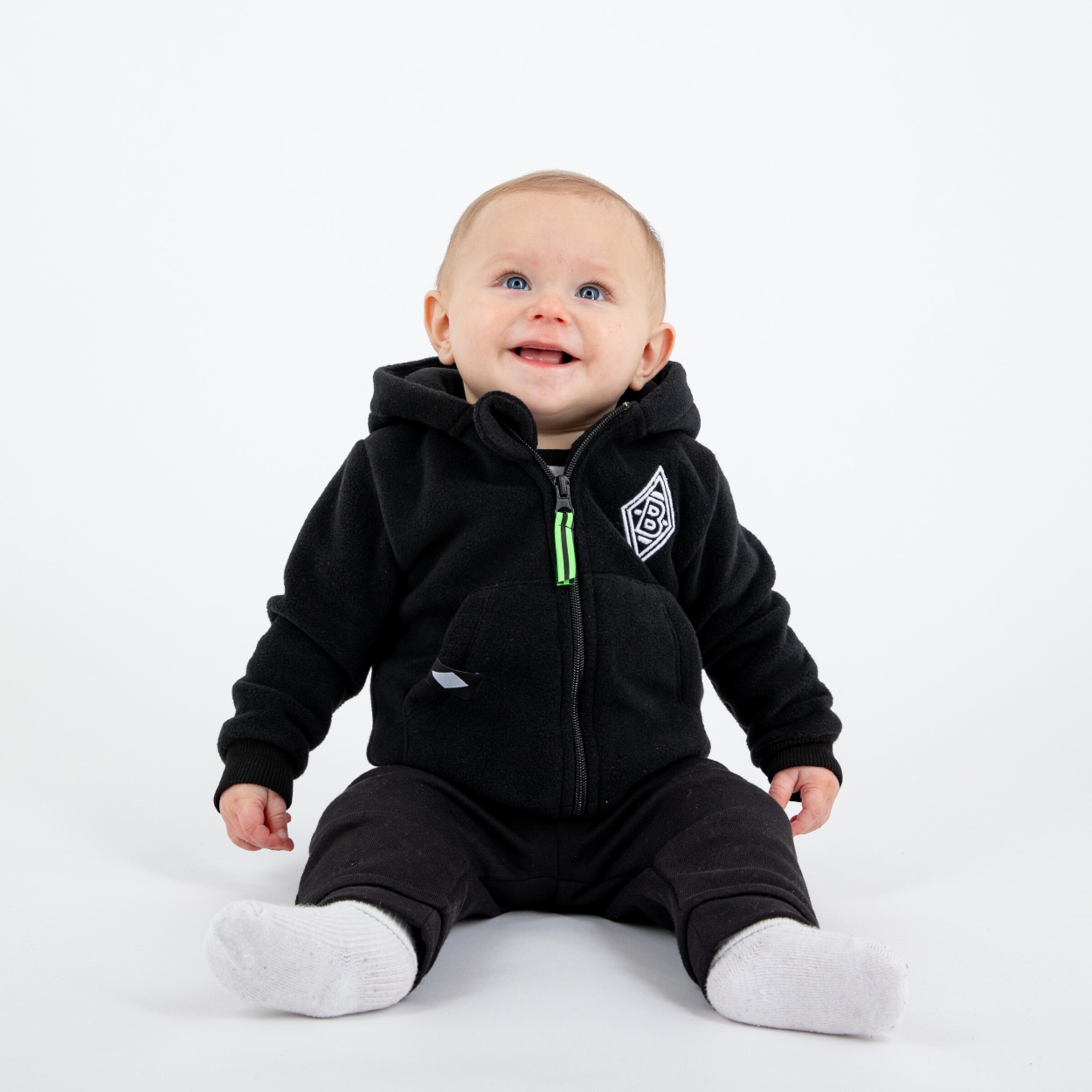 Ein lächelndes Baby sitzt auf dem Boden trägt einen schwarzen Hoodie mit dem Logo von Borussia Mönchengladbach und schwarze Hosen.