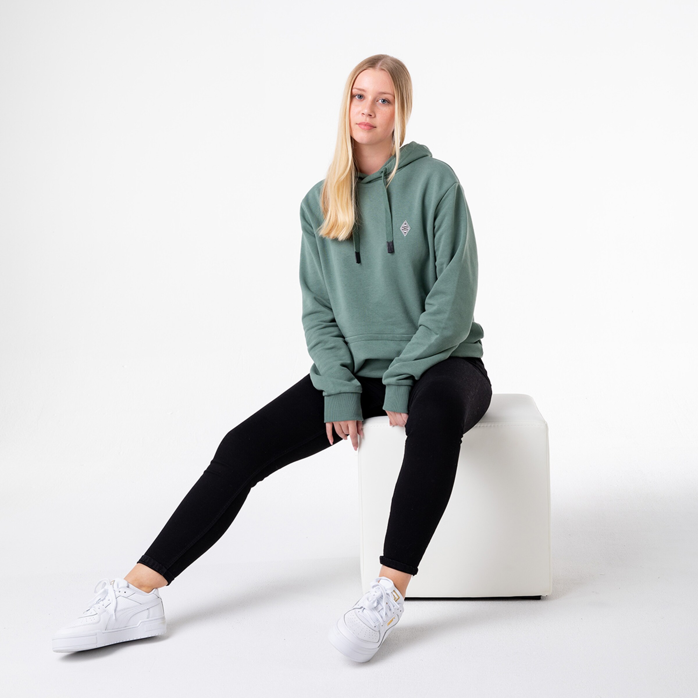 Dame mit langen blonden Haaren in einem grünen Hoodie und schwarzen Leggings sitzt auf einem weißen Würfel.