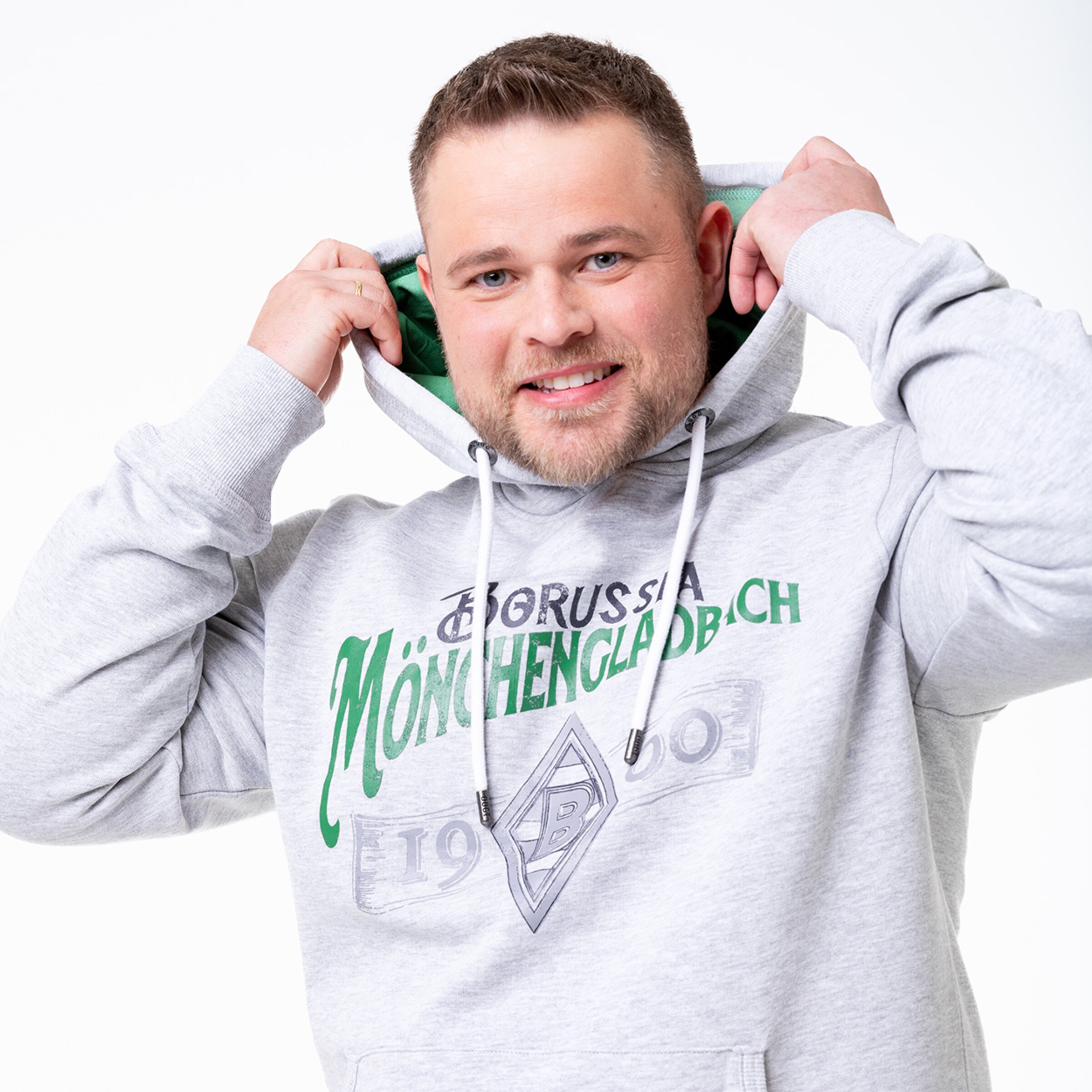 Ein Mann lächelt und zieht die Kapuze eines grauen Hoodies mit Borussia Mönchengladbach-Logo über seinen Kopf.