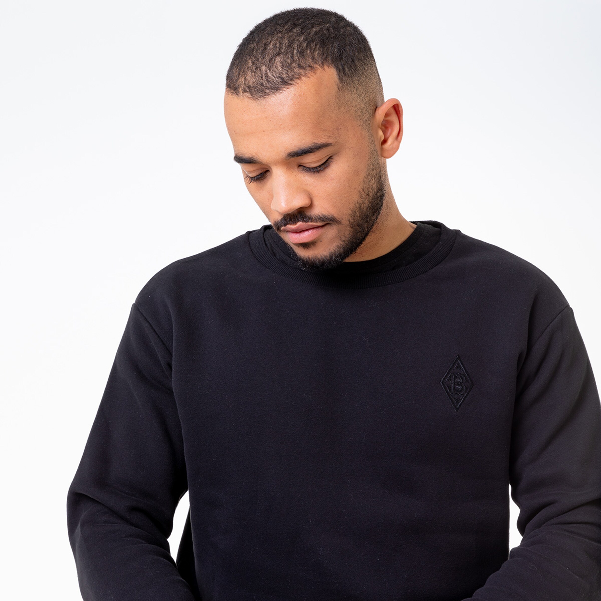Man wearing a black sweatshirt with an embroidered logo on the chest looking down.