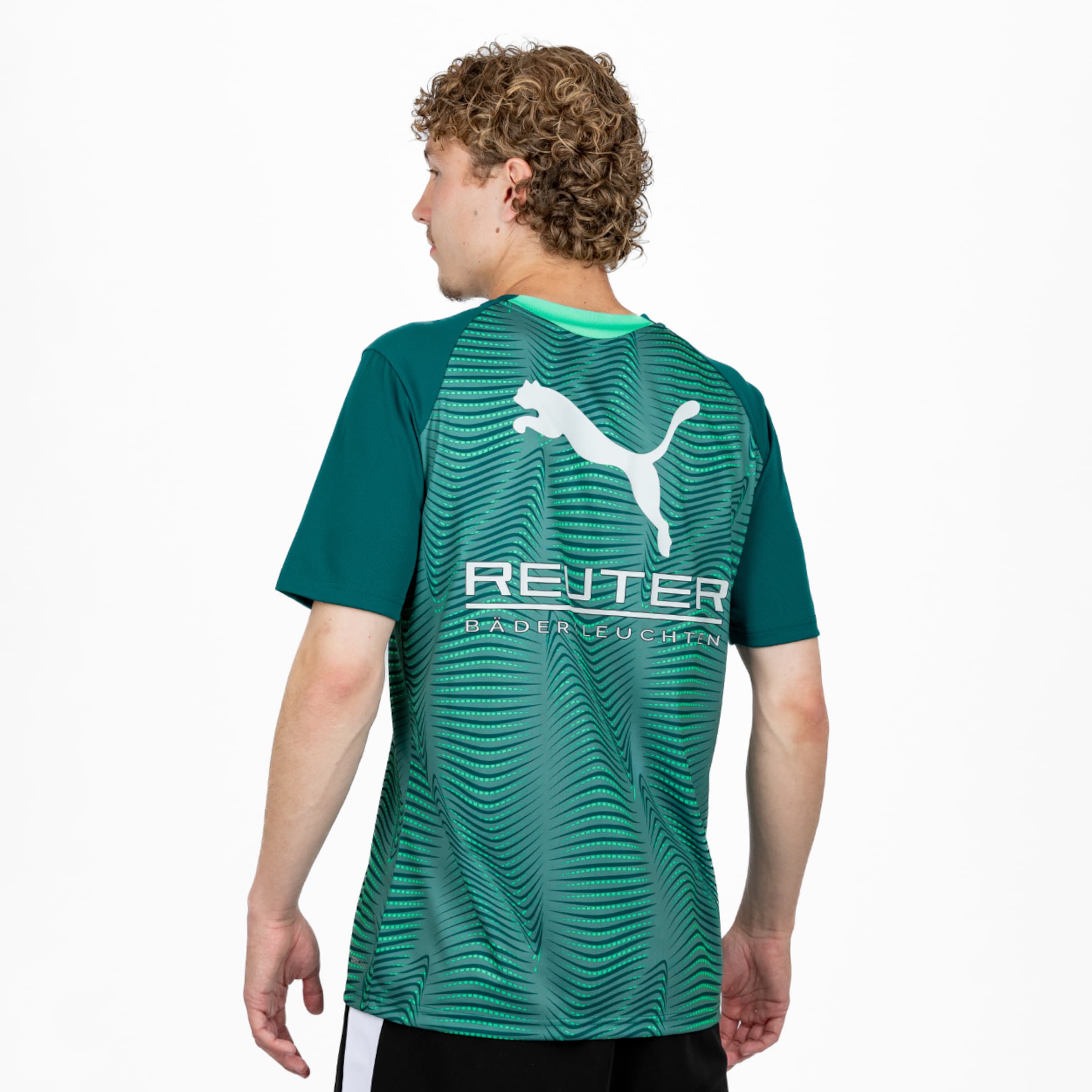 Ein Mann von hinten, trägt ein grün gemustertes PUMA Sporttrikot mit dem Schriftzug 'REUTER BÄDERLEUCHTEN'. Er hat lockiges Haar und blickt nach links vor weißem Hintergrund.