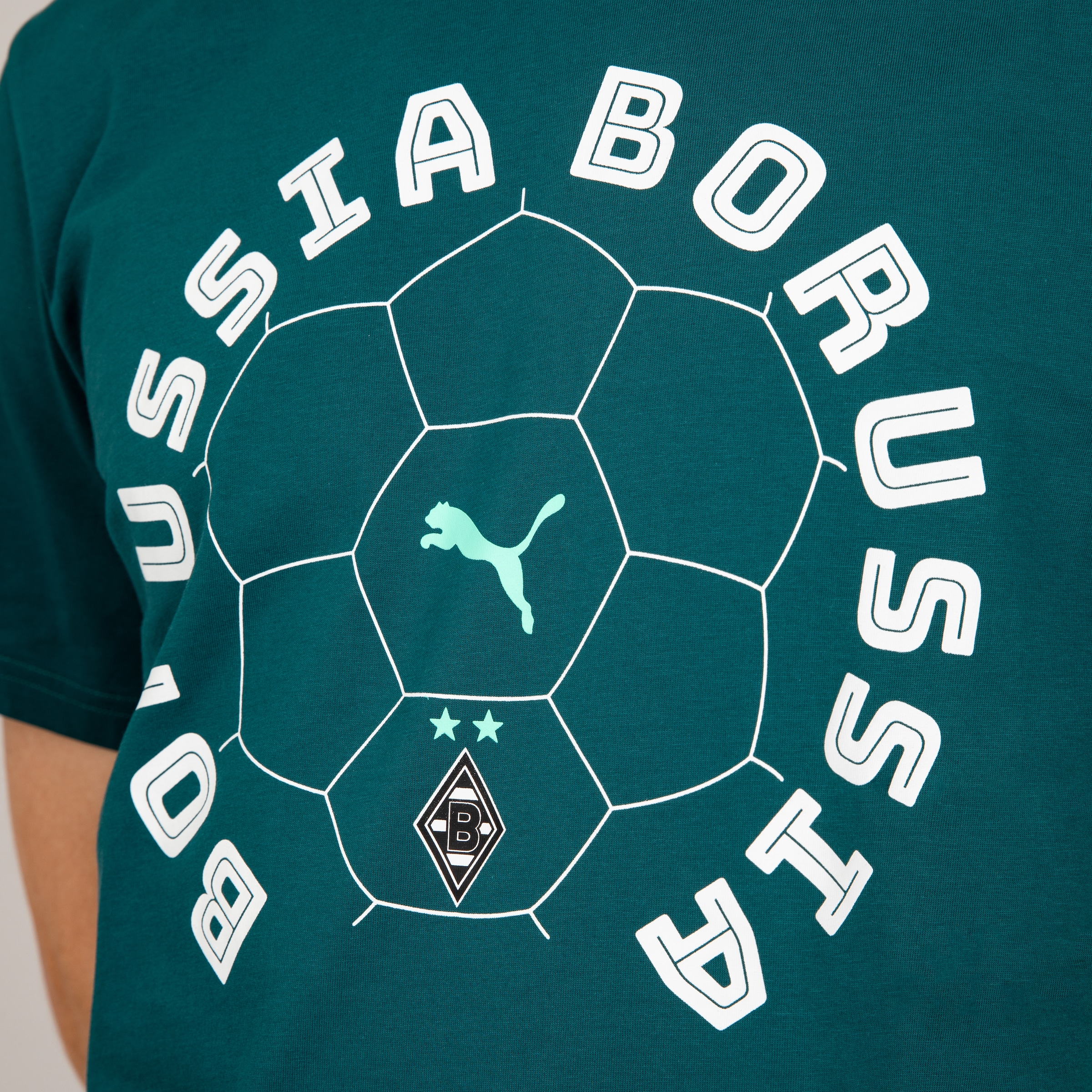 Nahaufnahme eines petrolfarbenen T-Shirts, auf dem in einem großen weißen Halbkreis die Worte "BORUSSIA" und "MONCHENGLADBACH" stehen. In der Mitte ist ein Fußballumriss mit dem Puma-Logo und dem Borussia Mönchengladbach-Wappen zu sehen.