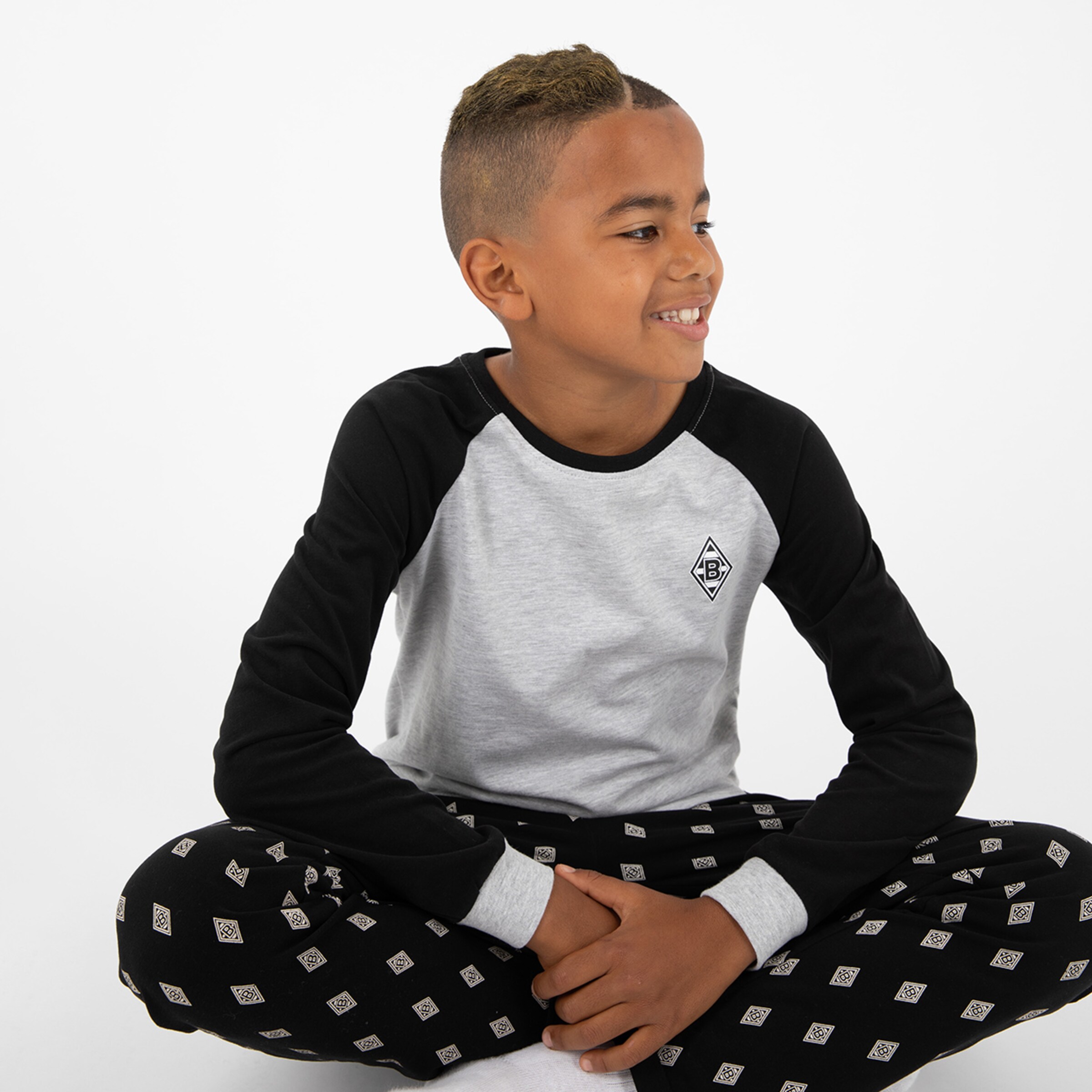 Junge sitzt mit gekreuzten Beinen trägt grauen T-Shirt mit schwarzem Ärmel und schwarze Hose mit Muster.