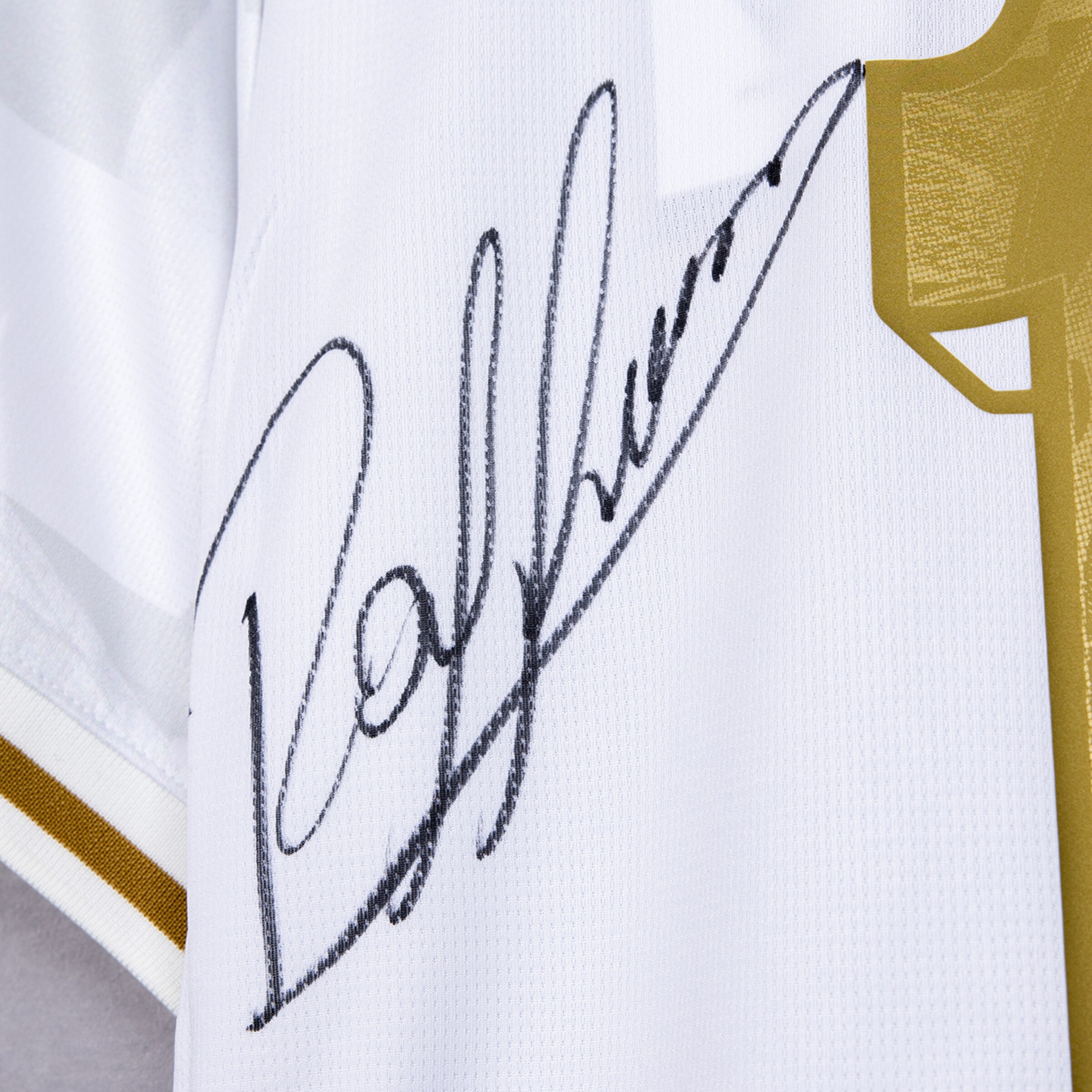 Nahaufnahme einer schwarzen handschriftlichen Signatur auf einem weißen Trikot mit goldenen Details.
