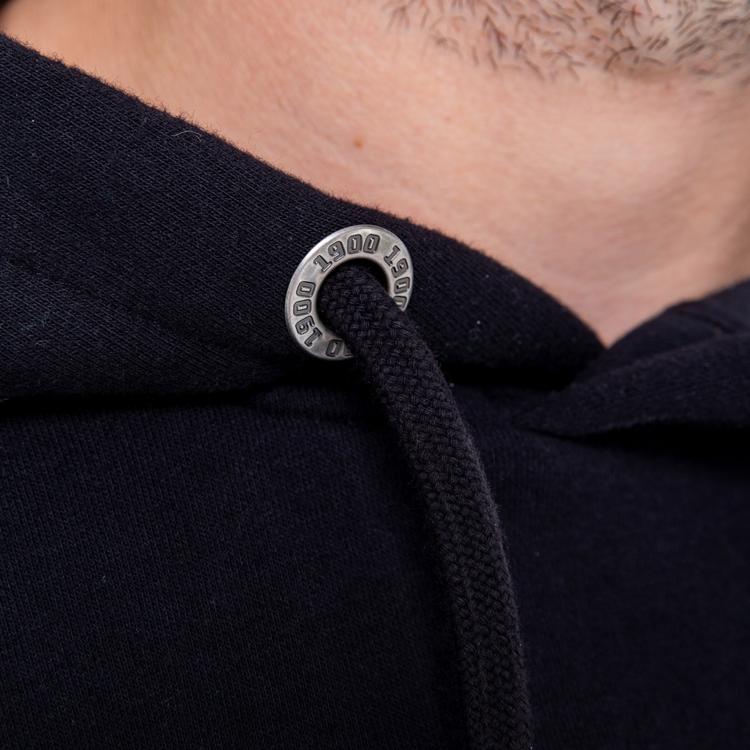 Detailaufnahme des Kordelstopper einer schwarzen Kapuzenjacke mit der Prägung 1900.