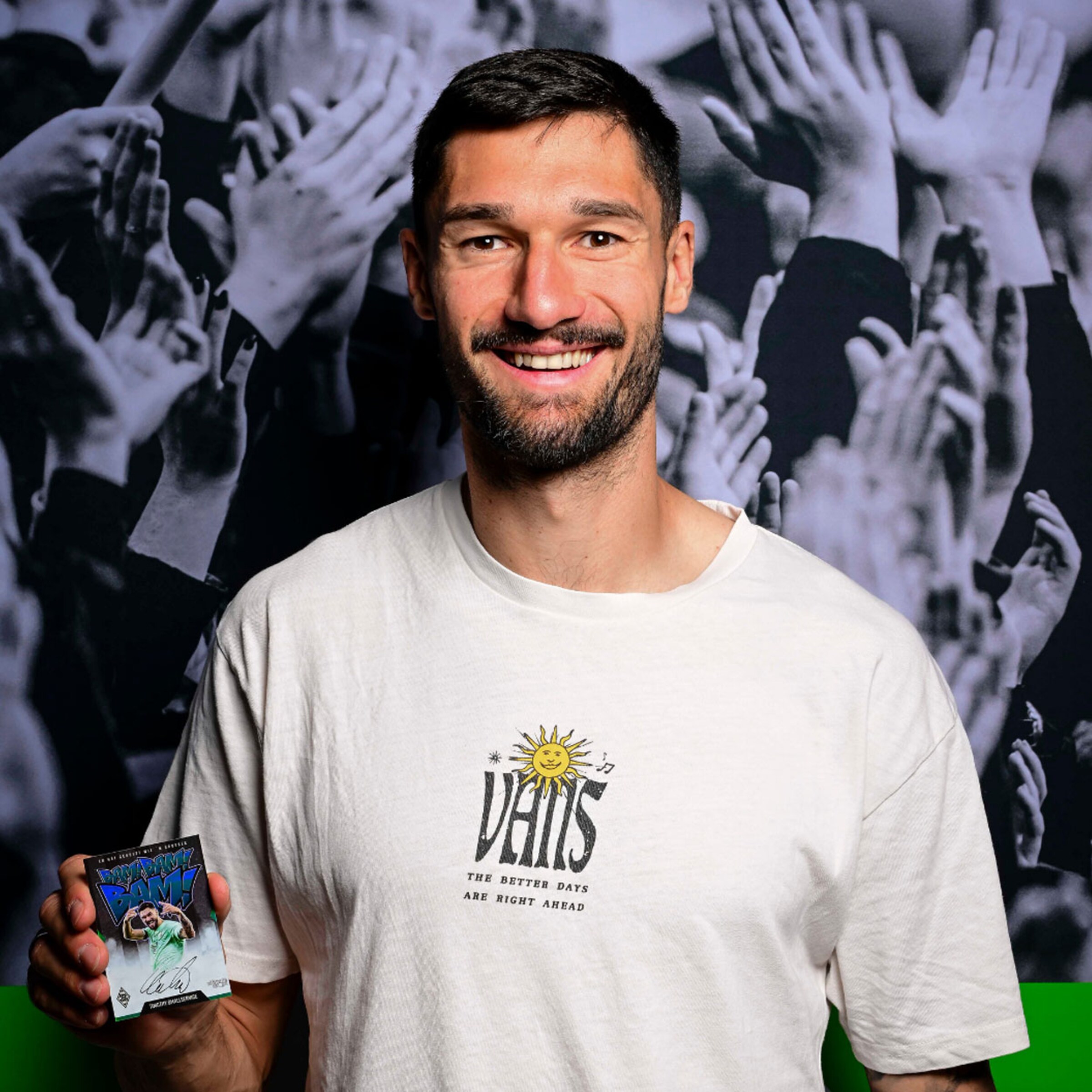 Ein Spieler von Borussia Mönchengladbach hält eine Sammelkarte in der Hand und lächelt vor einer jubelnden Menge.