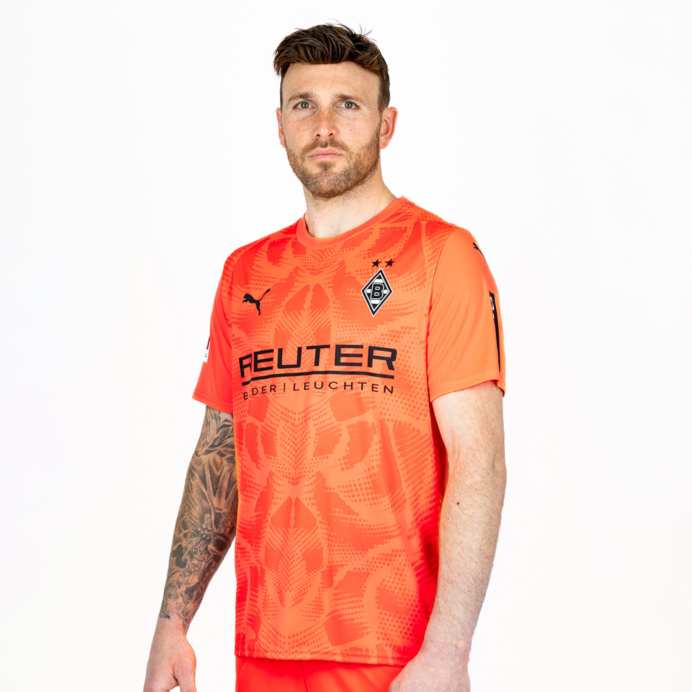 Borussia Mönchengladbach player in an orange Puma goalkeeper jersey with REUTER sponsor and arm tattoos, against a white background.