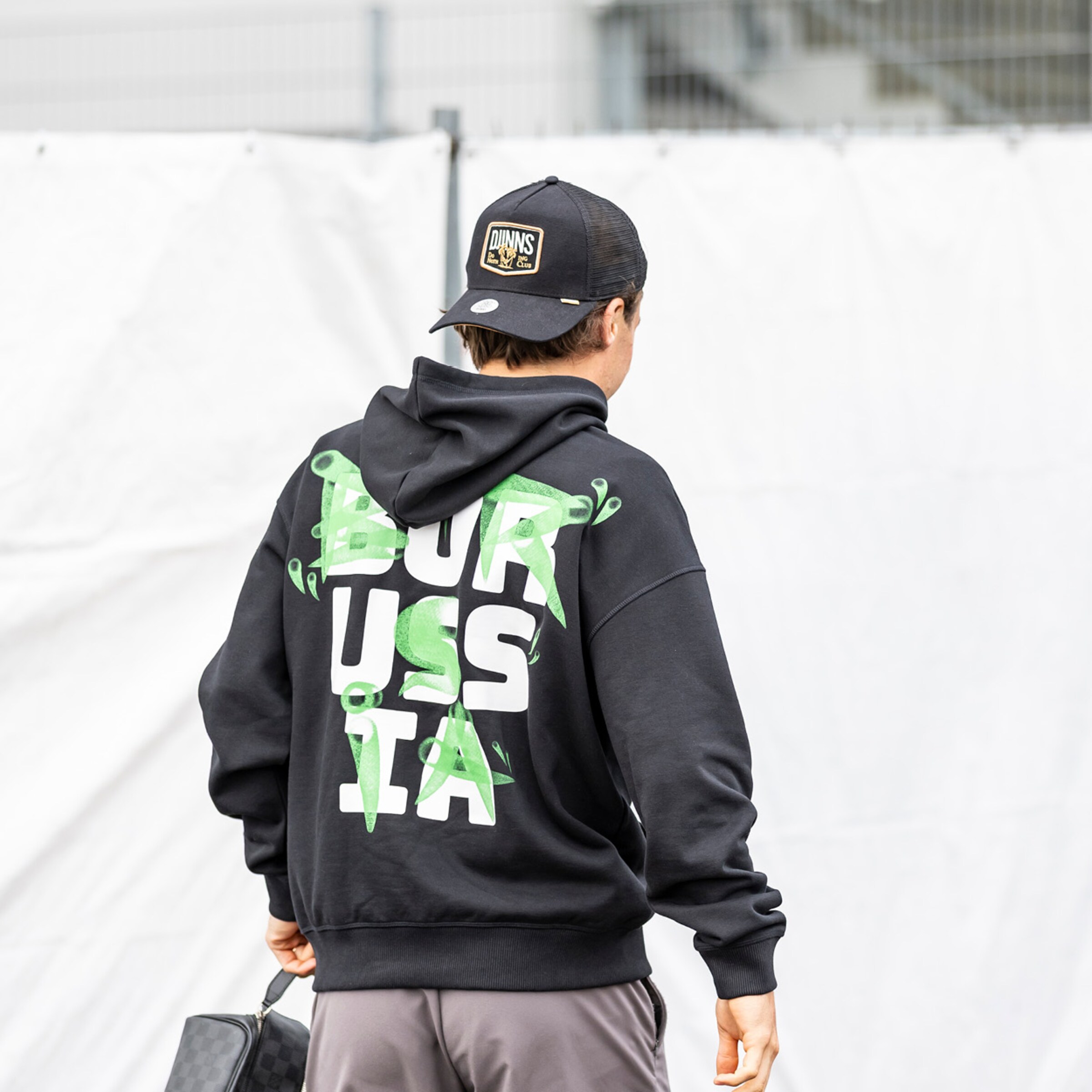 Rückansicht eines Mannes in schwarzem Hoodie mit grün-weißem „BORUSSIA“-Schriftzug und schwarzer Cap vor weißem Hintergrund.