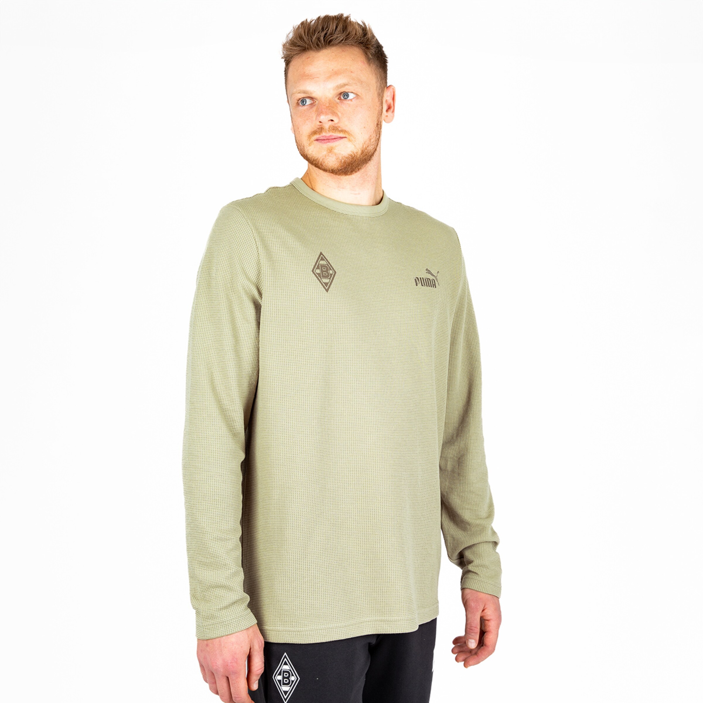 Ein Mann in einem langen Shirt steht seitlich trägt ein olivfarbenes Shirt mit Logo der Marke Puma und Borussia Mönchengladbach.