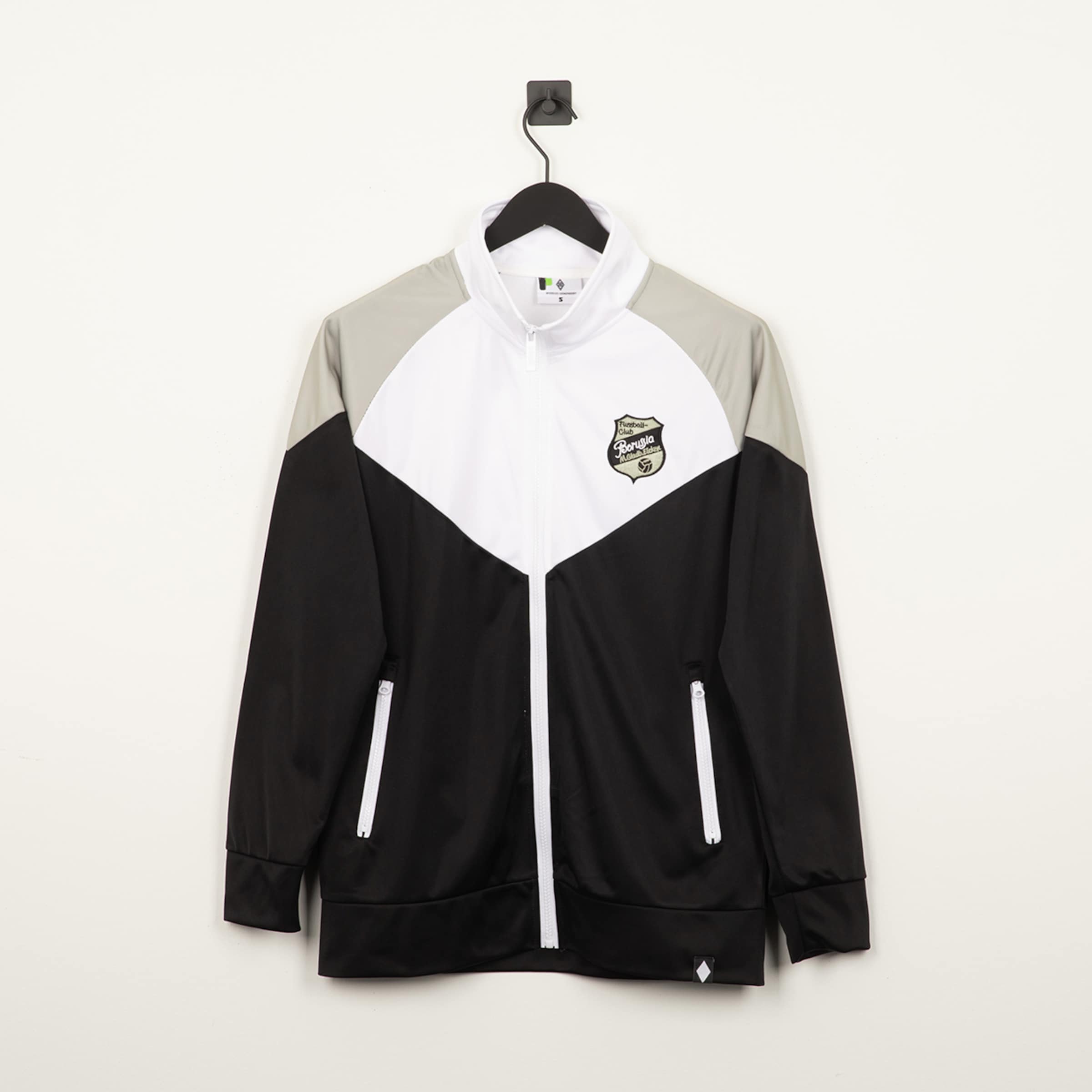 Schwarz-weiß-graue Trainingsjacke mit Borussia-Wappen, durchgehendem Reißverschluss und V-Design, hängend vor weißem Hintergrund.