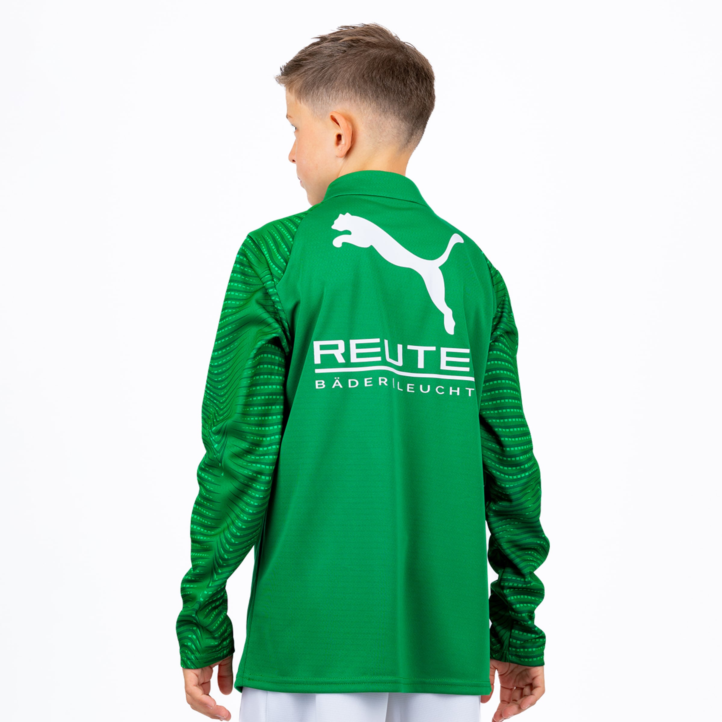 Ein Junge von hinten trägt ein grünes Langarm-Sportshirt mit einem weißen Puma-Logo und dem Schriftzug „REUTE BÄDER LEUCHT“ auf dem Rücken. Die Ärmel sind mit einem Streifenmuster versehen. Der Hintergrund ist weiß.