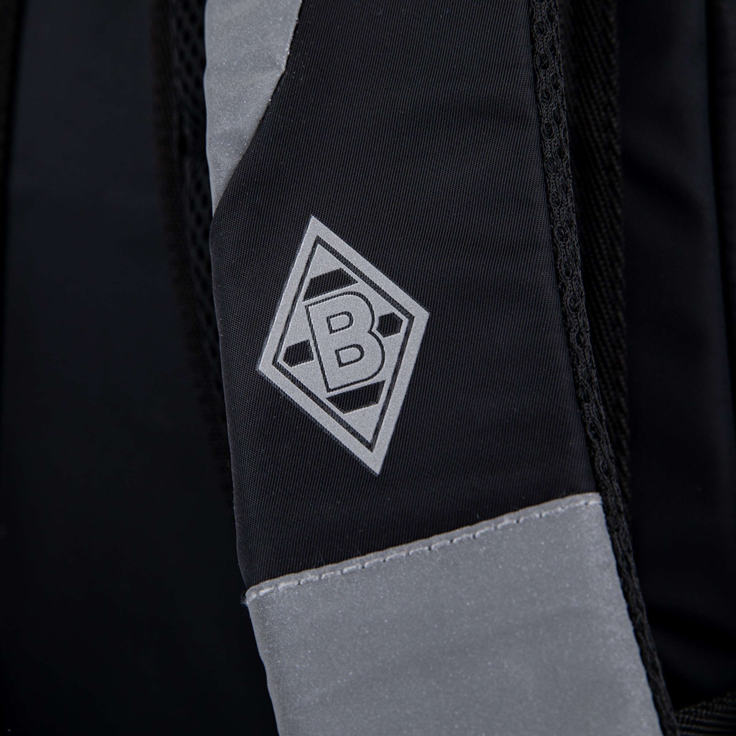 Close-up of a black fabric, likely a backpack, featuring a grey, reflective Borussia Mönchengladbach logo. The strap has grey reflective strips and a textured weave.