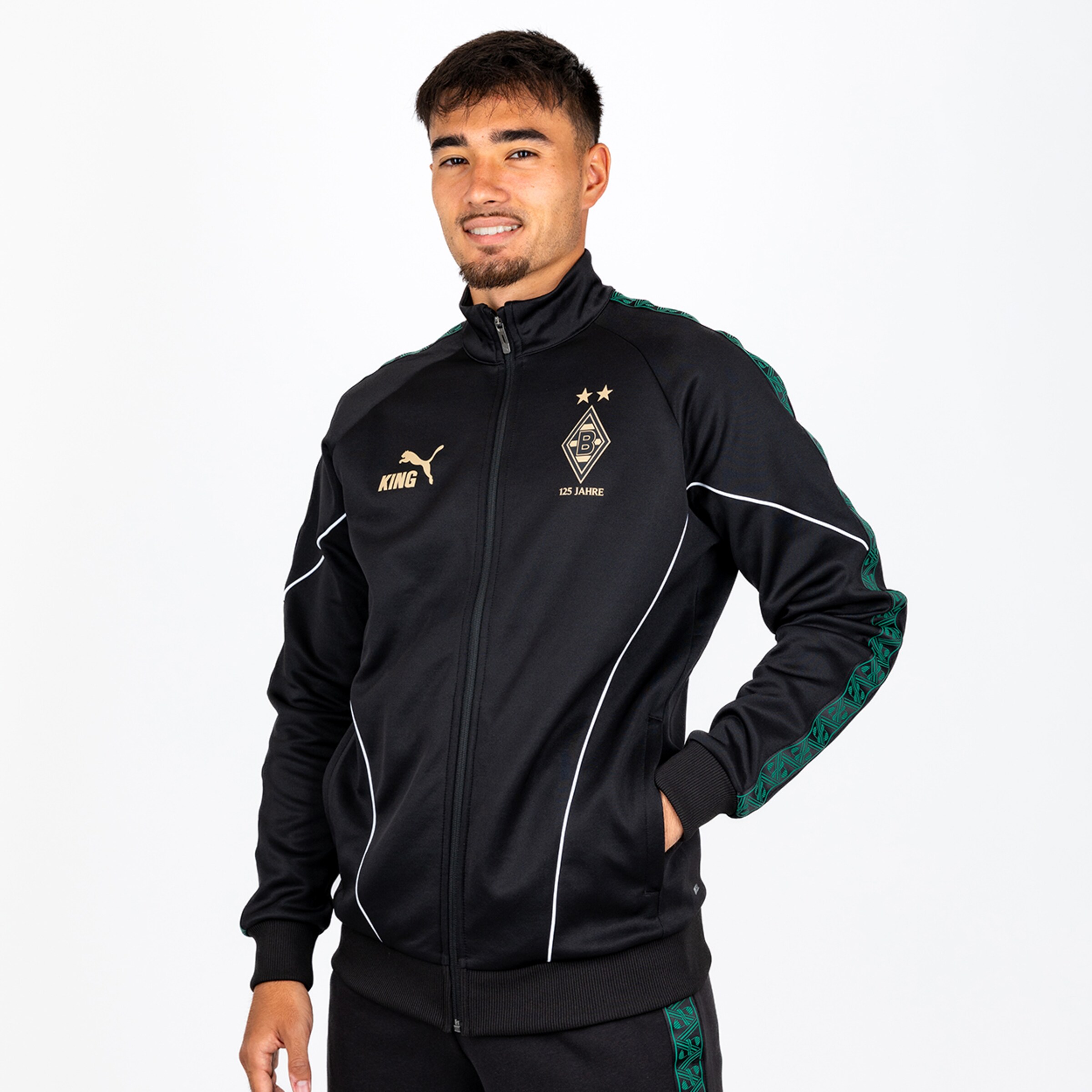 Mann in schwarzer Puma Borussia Mönchengladbach Jacke mit goldenen Logos, 125-Jahre-Jubiläumswappen und grünen Mustern an den Ärmeln.