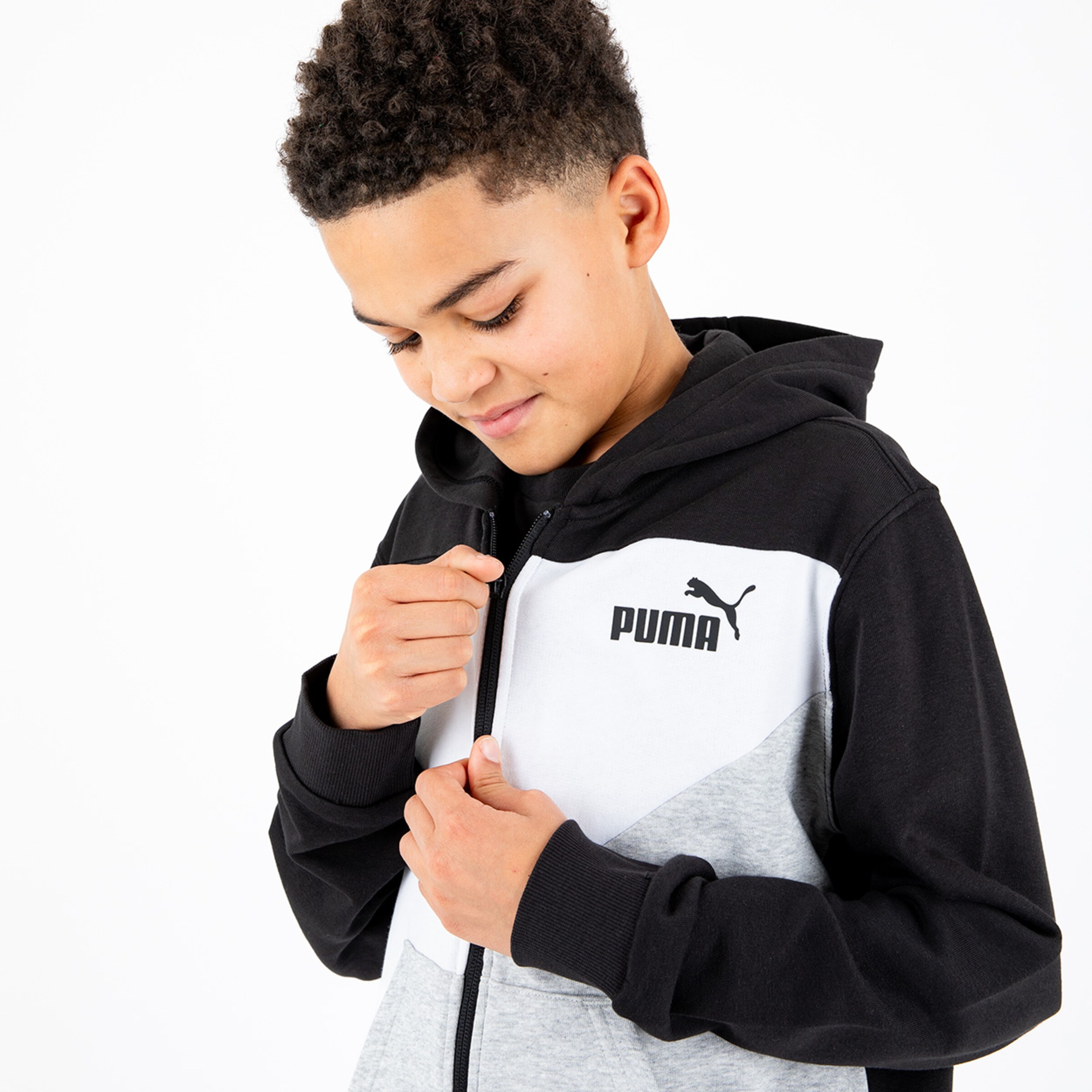 A boy wearing a black gray and white Puma hoodie is zipping it up looking thoughtful and smiling slightly.