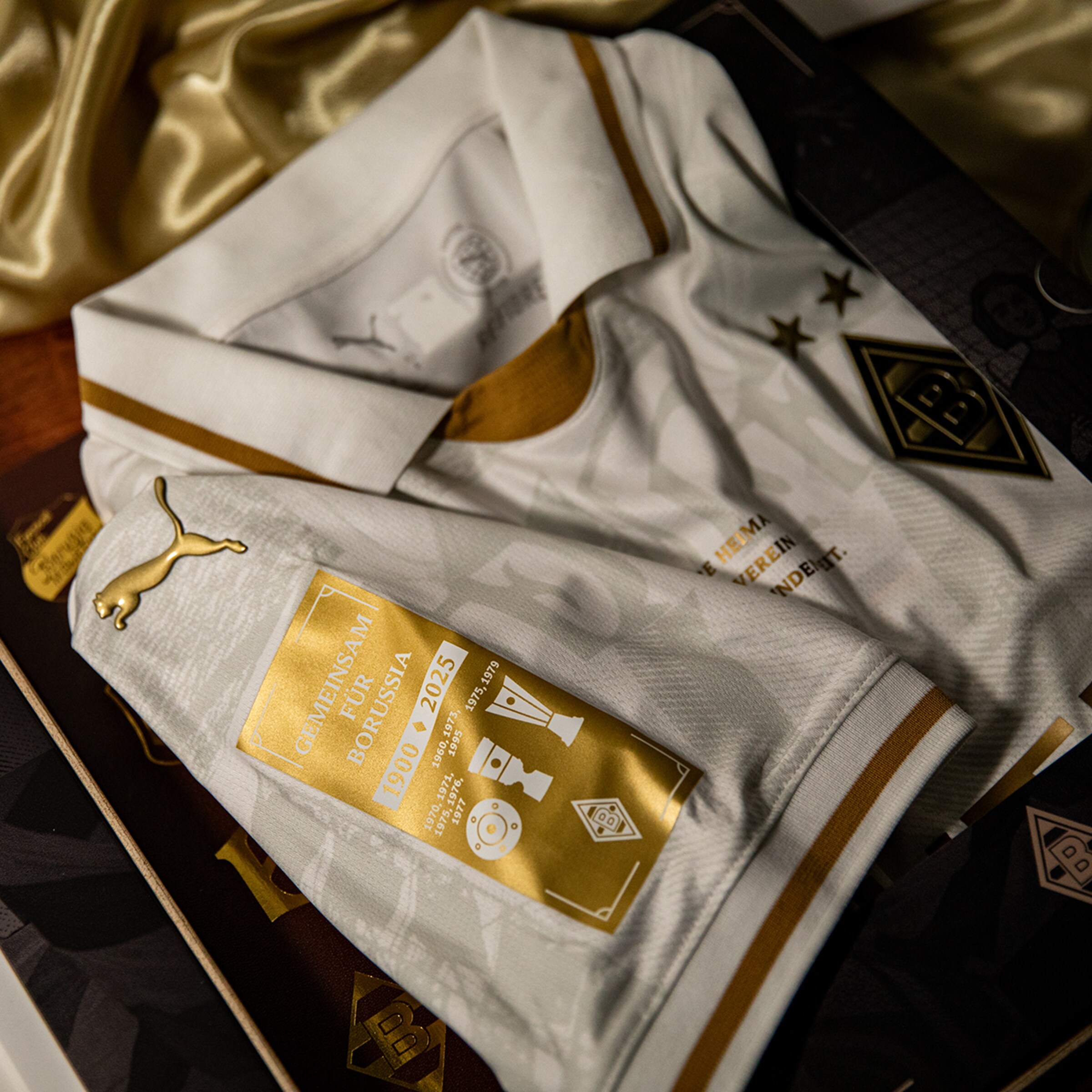 Weißes Borussia-Mönchengladbach-Jubiläumstrikot mit goldenen Details, Vereinswappen und Aufnäher mit Titelerfolgen der Jahre 1900 bis 2025.