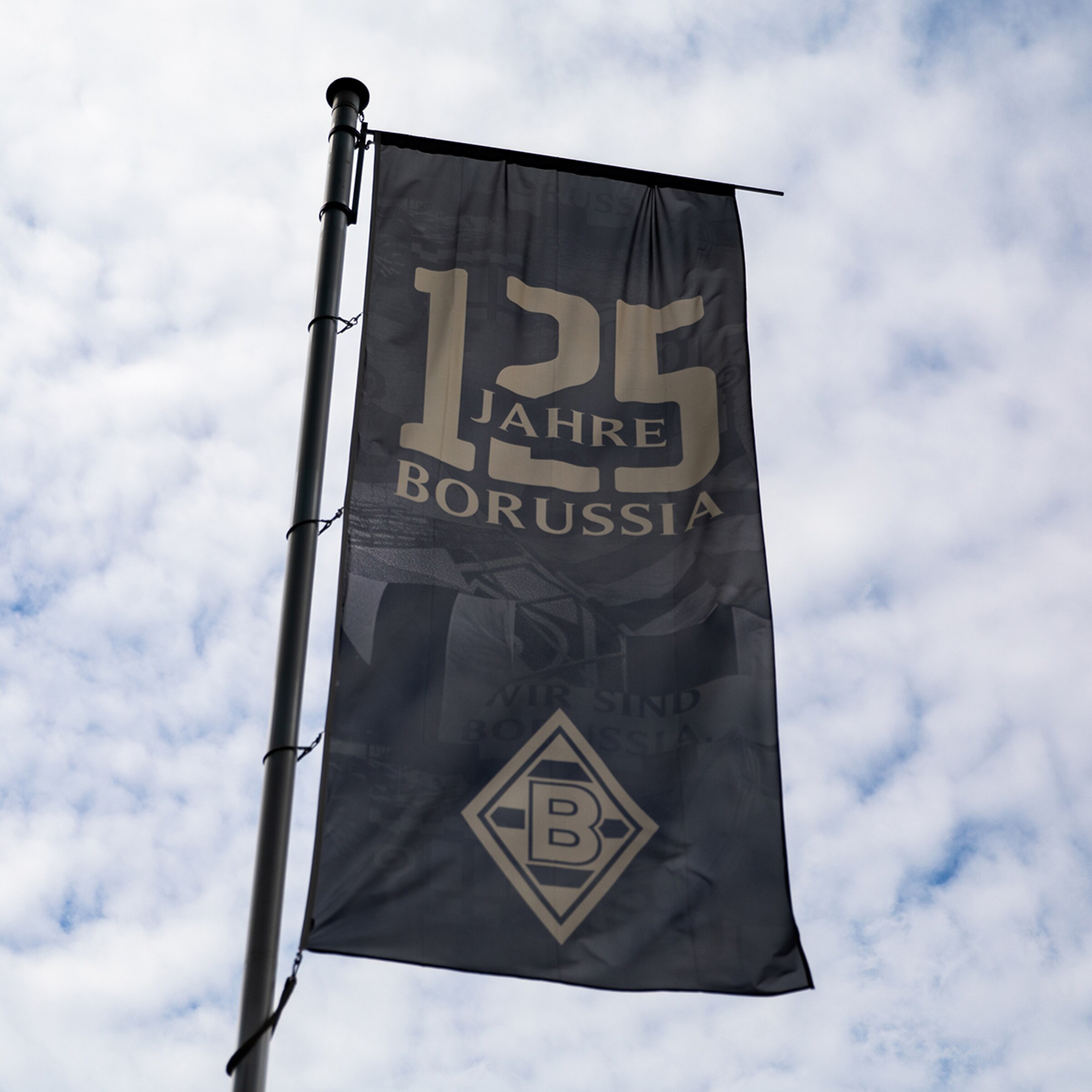 Dunkelgraue Fahne zum 125. Jubiläum von Borussia Mönchengladbach mit Logo und Aufschrift vor bewölktem Himmel.