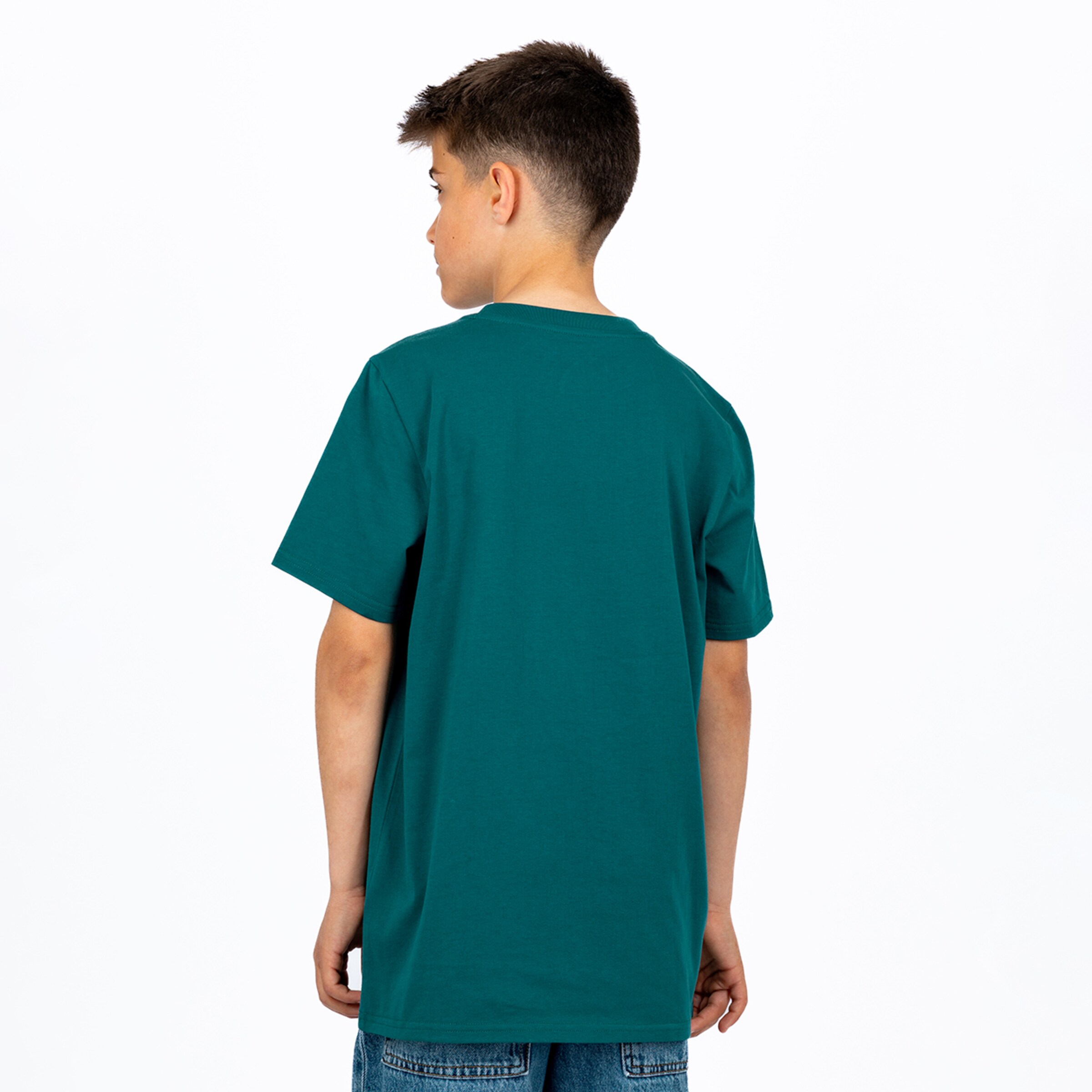 Rückenansicht eines jungen Jungen in einem schlichten petrolfarbenen Kurzarm-T-Shirt und blauen Jeans, leicht nach links gedreht vor weißem Hintergrund.