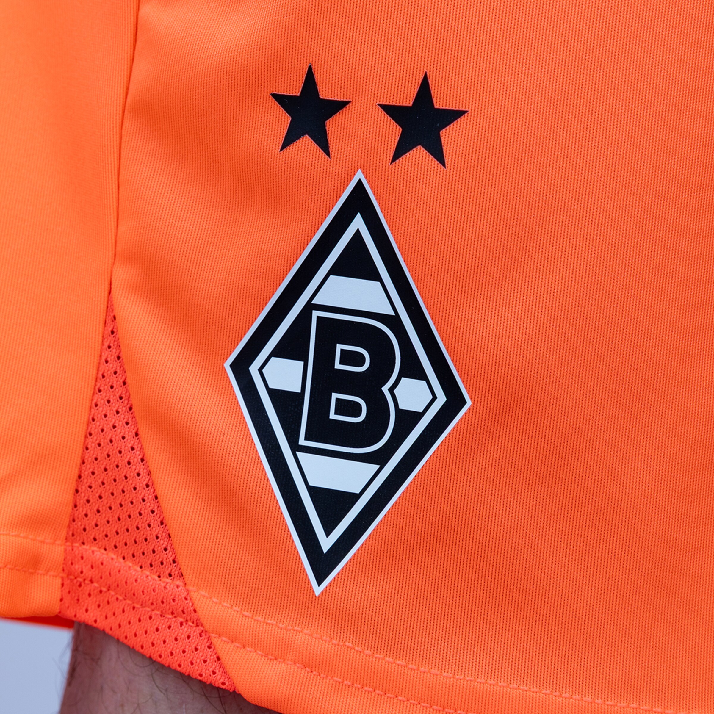 Vereinslogo von Borussia Mönchengladbach auf orangefarbenem Stoff: Eine schwarz-weiße Raute mit einem „B“ und zwei schwarzen Sternen darüber.