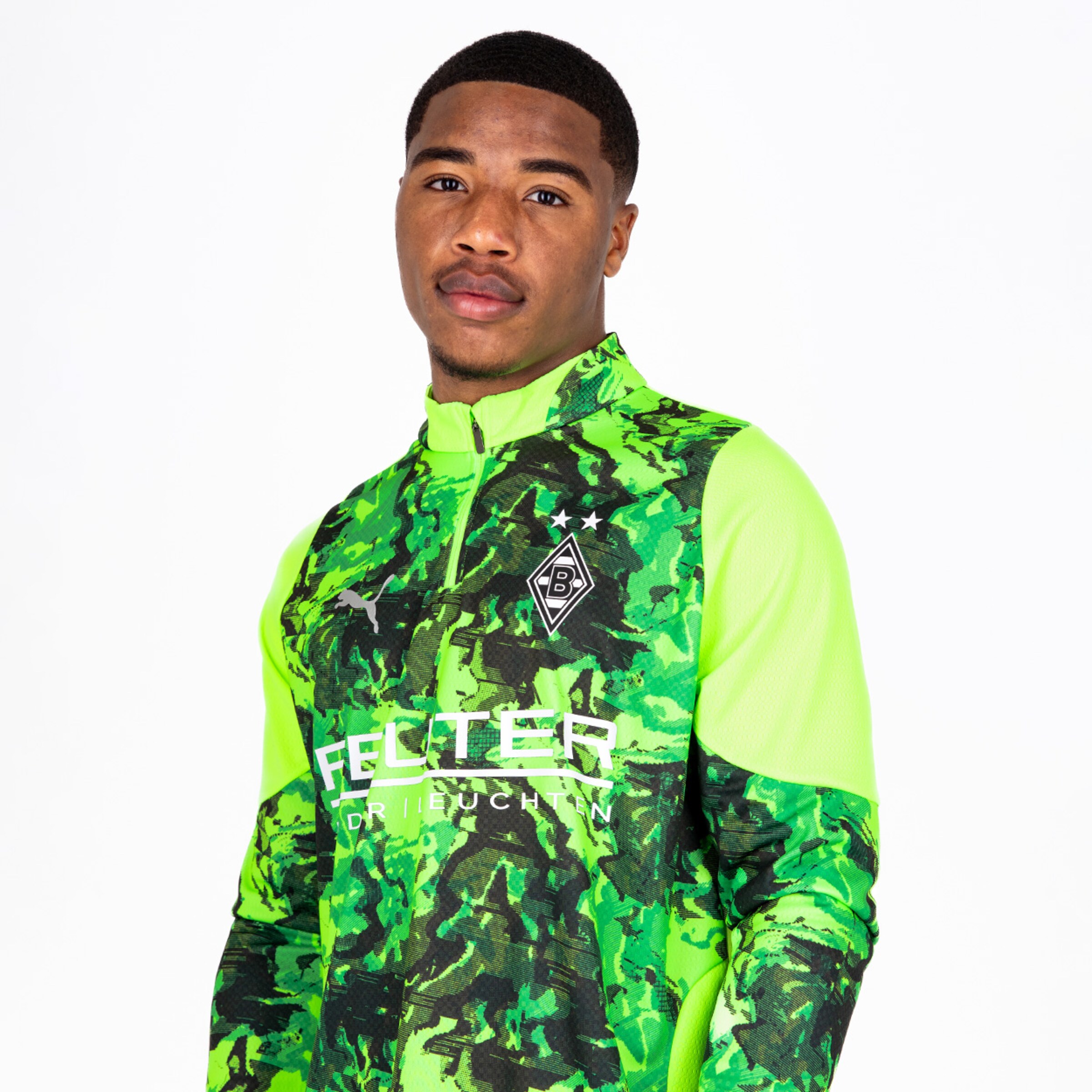 Ein junger Fußballspieler trägt ein grünes Trikot mit einem camouflagemuster und dem Logo von Borussia Mönchengladbach.