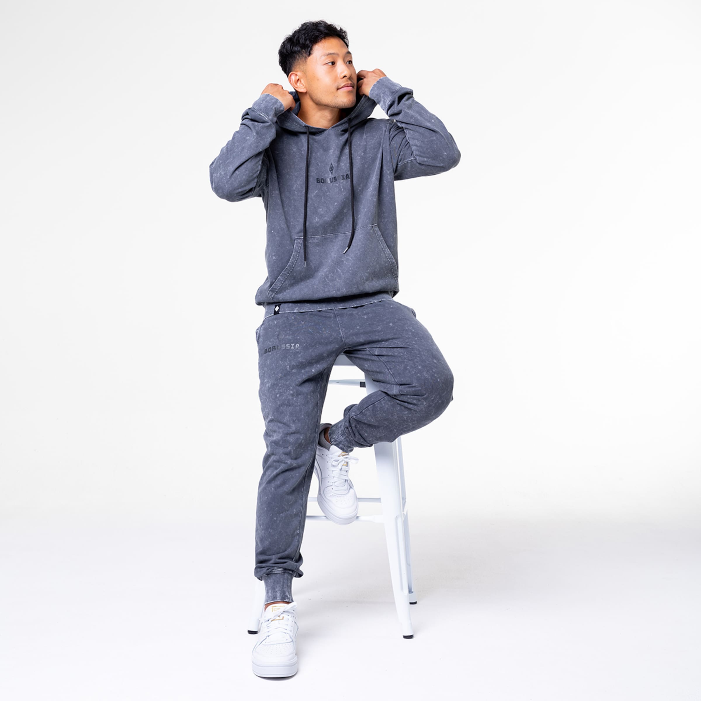 Mann sitzt auf einem Hocker in grauer Sportbekleidung mit Kapuzenpullover und Jogginghose.