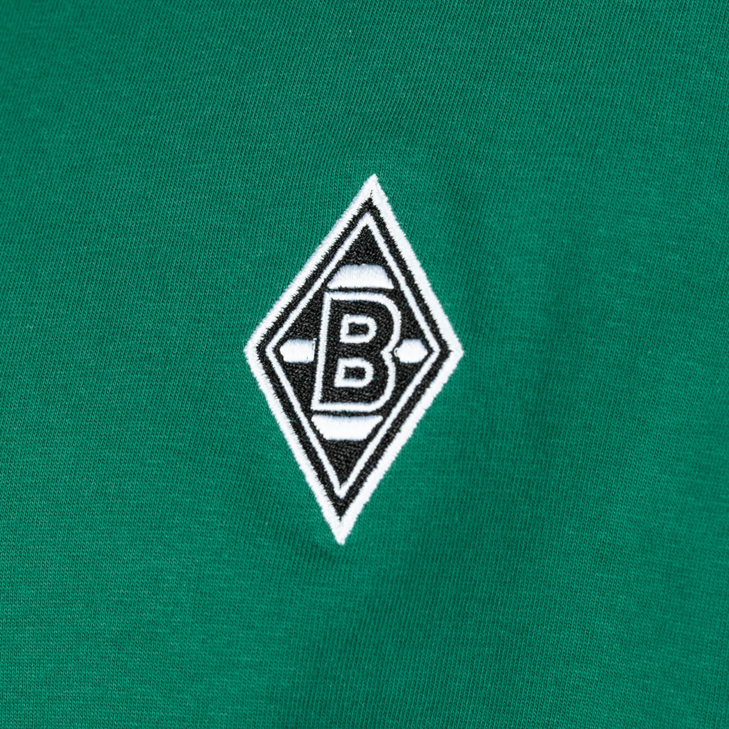 Nahaufnahme eines gestickten Logos auf grünem Stoff. Das Logo ist rautenförmig mit einem weißen Rand, einer schwarzen Fläche und einem stilisierten weißen Buchstaben 'B' in der Mitte. Es handelt sich um das Emblem von Borussia Mönchengladbach.